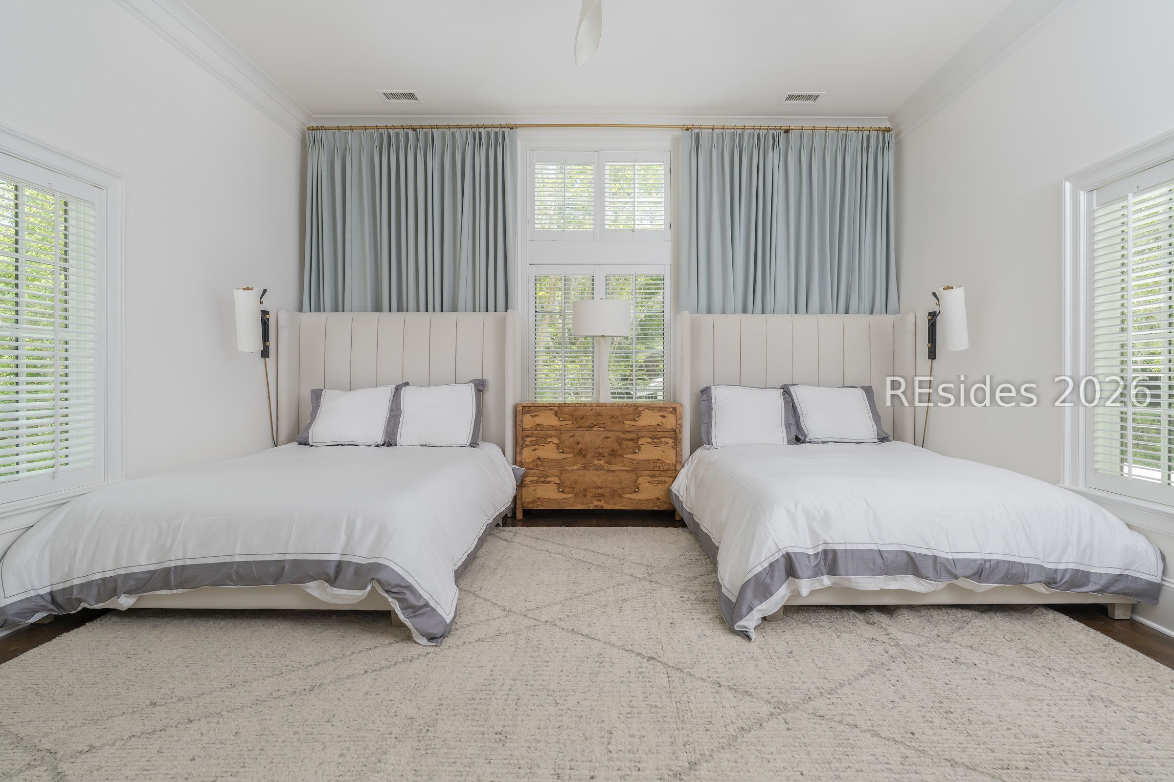 Bedroom with two white beds, shuttered windows, gray curtains, and a wood dresser between them.