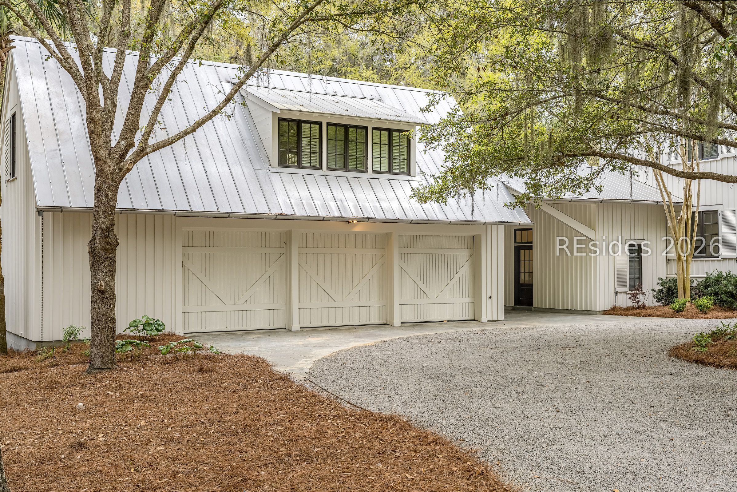 Light-colored home with three carriage-style garage doors beneath a metal roof, framed by mature trees.