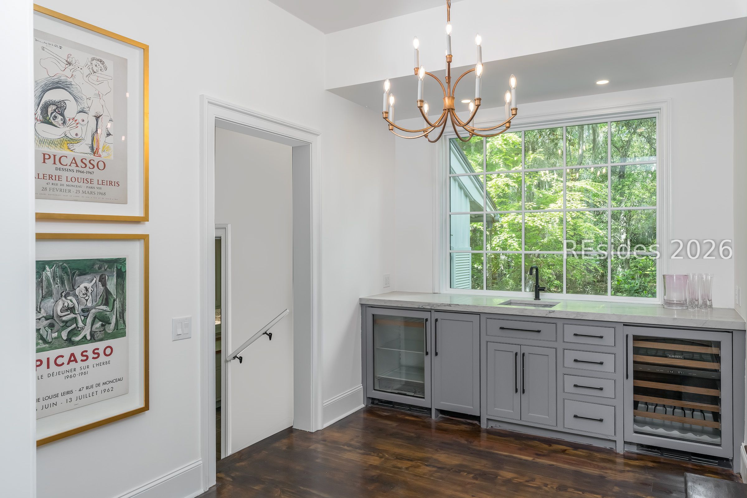 Wet bar with gray cabinetry, wine refrigerators, and a sink beneath a large window overlooking greenery.