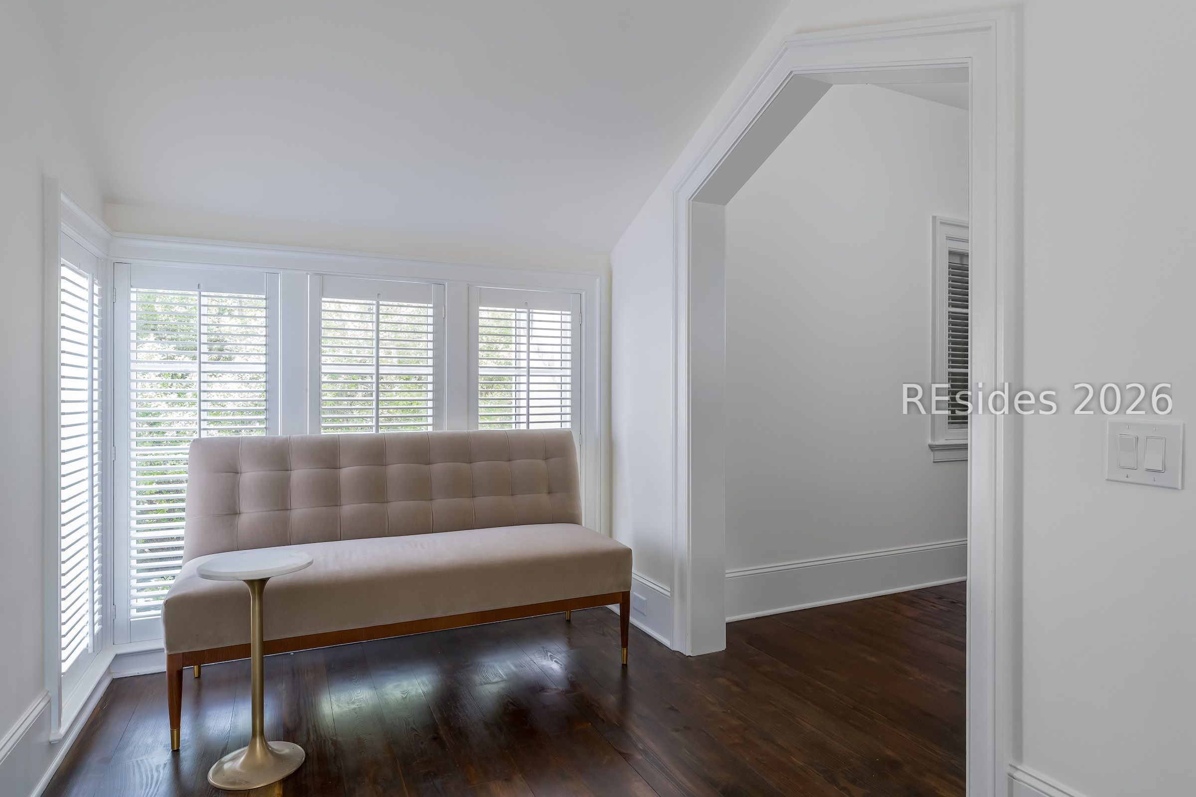 Sitting area with a beige settee, shuttered windows, and dark wood floors.