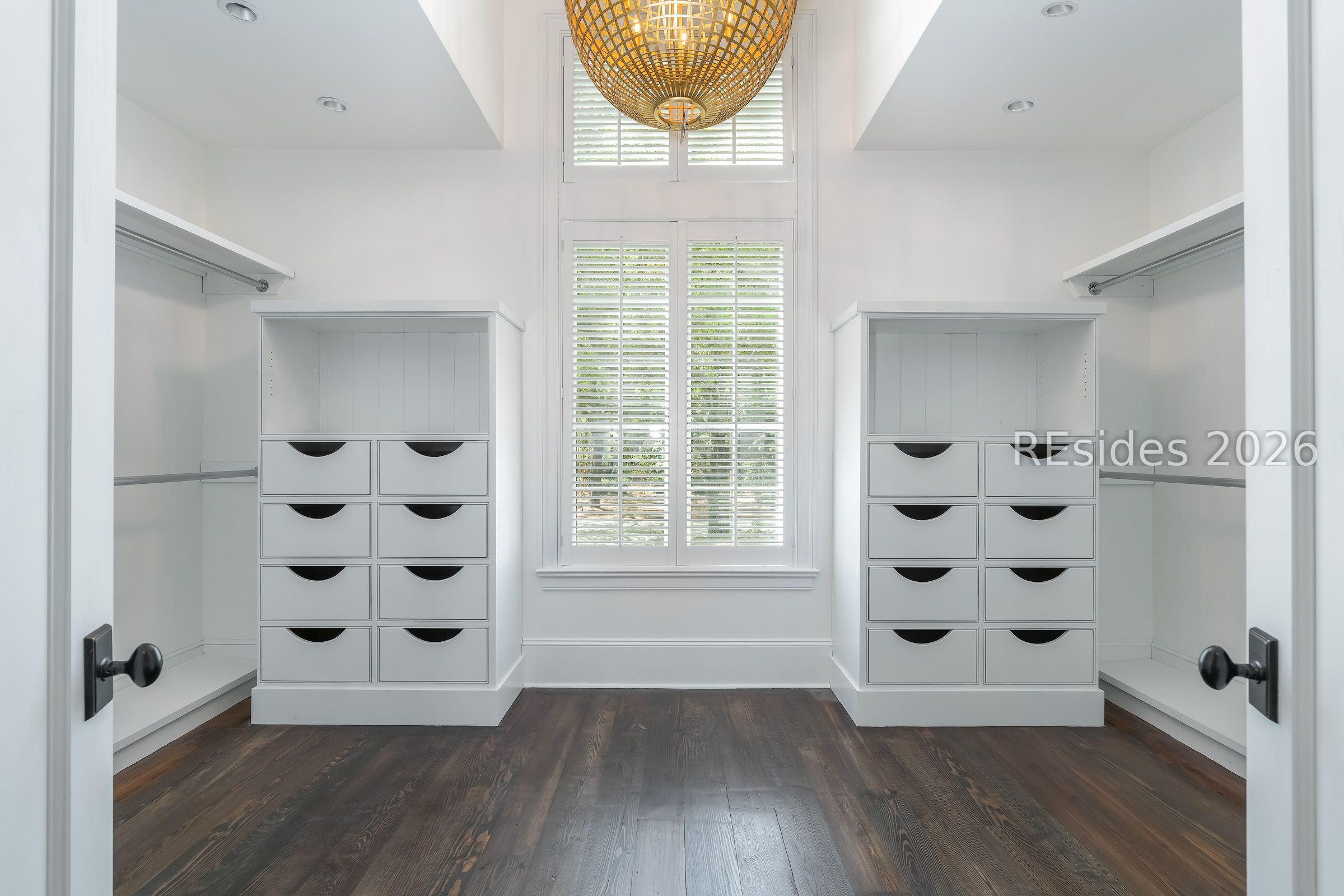 Walk-in closet with symmetrical white drawers, hanging rods, tall shuttered window, and dark wood flooring.