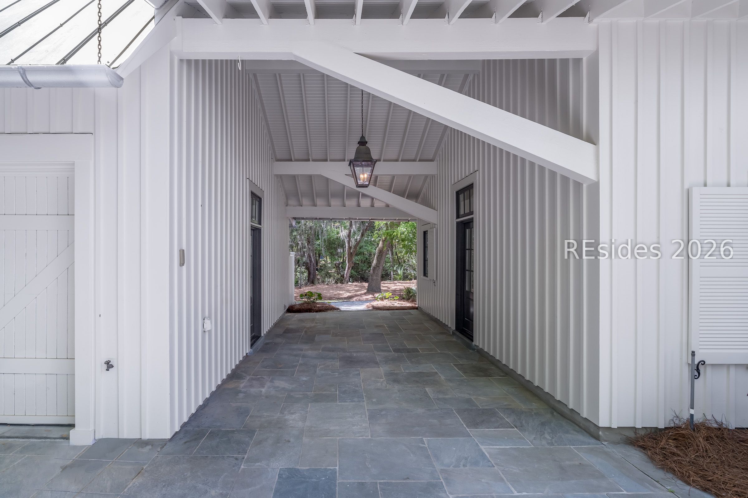 Covered entry passage with white walls, gray stone tile, exposed beams, and a view of trees.