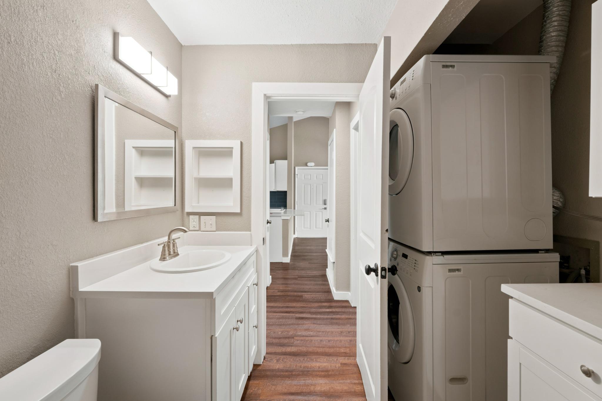 Bathroom vanity and sink beside a stacked washer and dryer, with a wood-look hallway beyond.