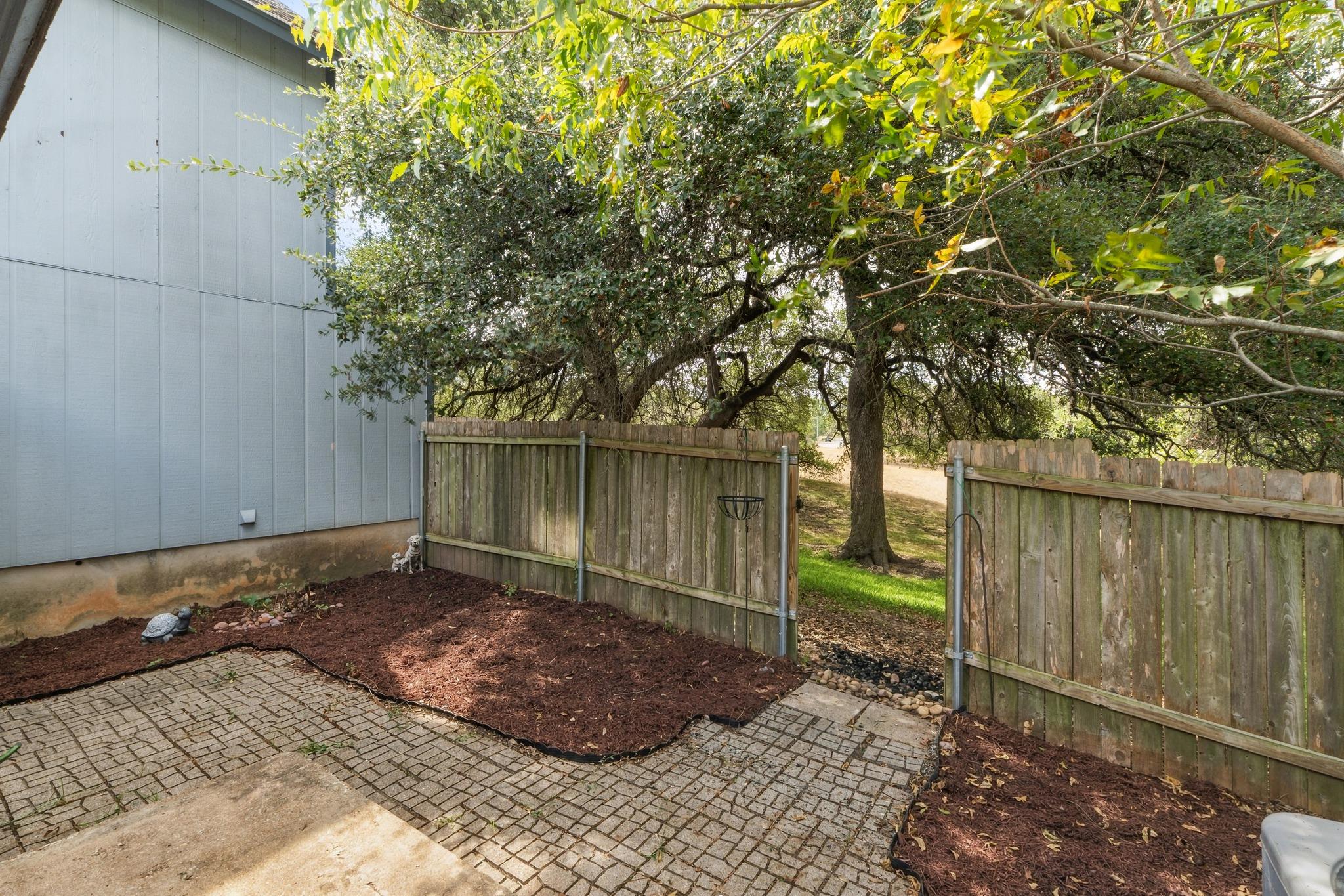 Fenced backyard patio with brick pavers, mulched garden beds, and mature shade trees.