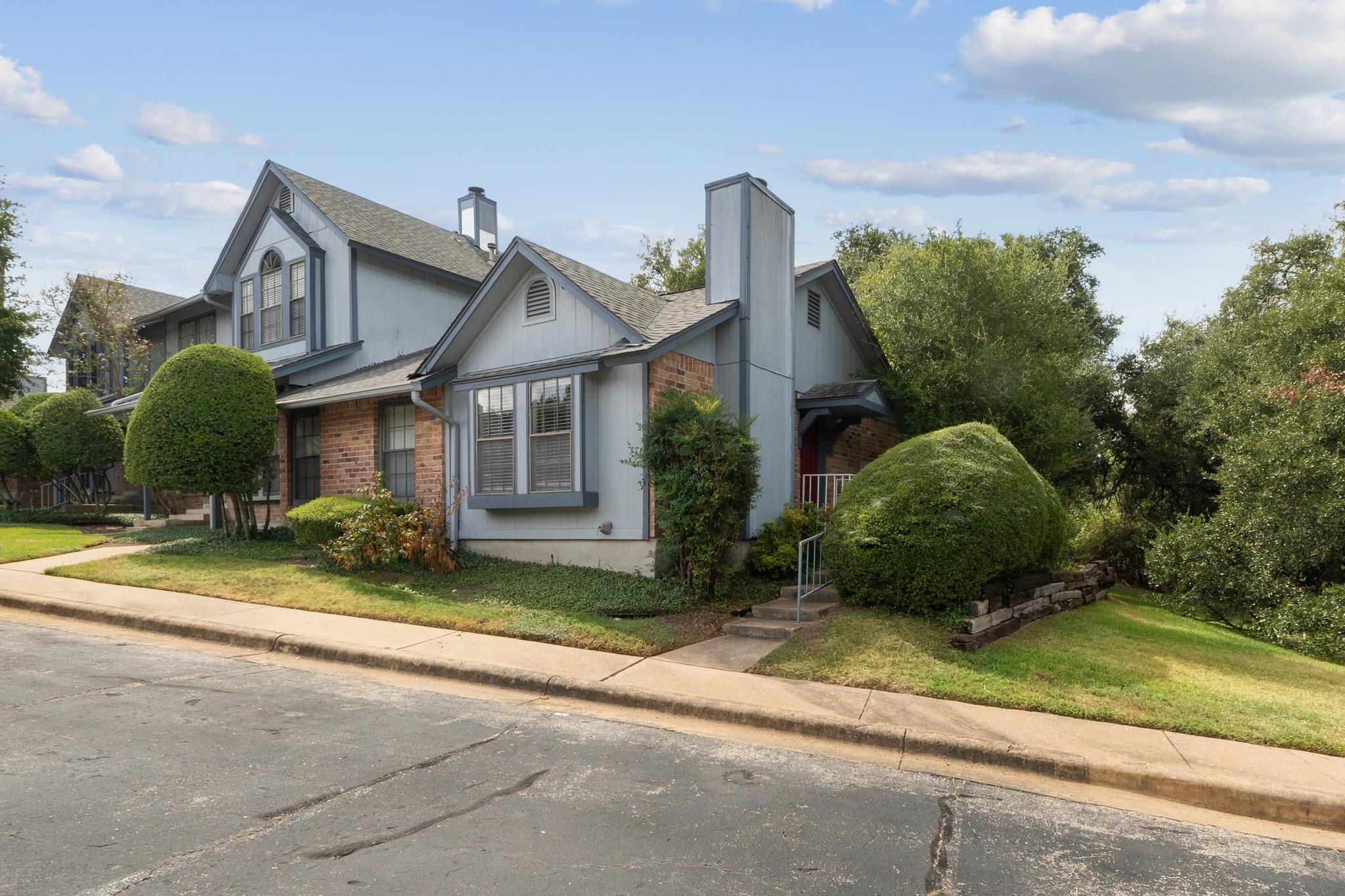 Two-story townhouse with gray siding, brick accents, gabled roofs, and neatly trimmed shrubs.