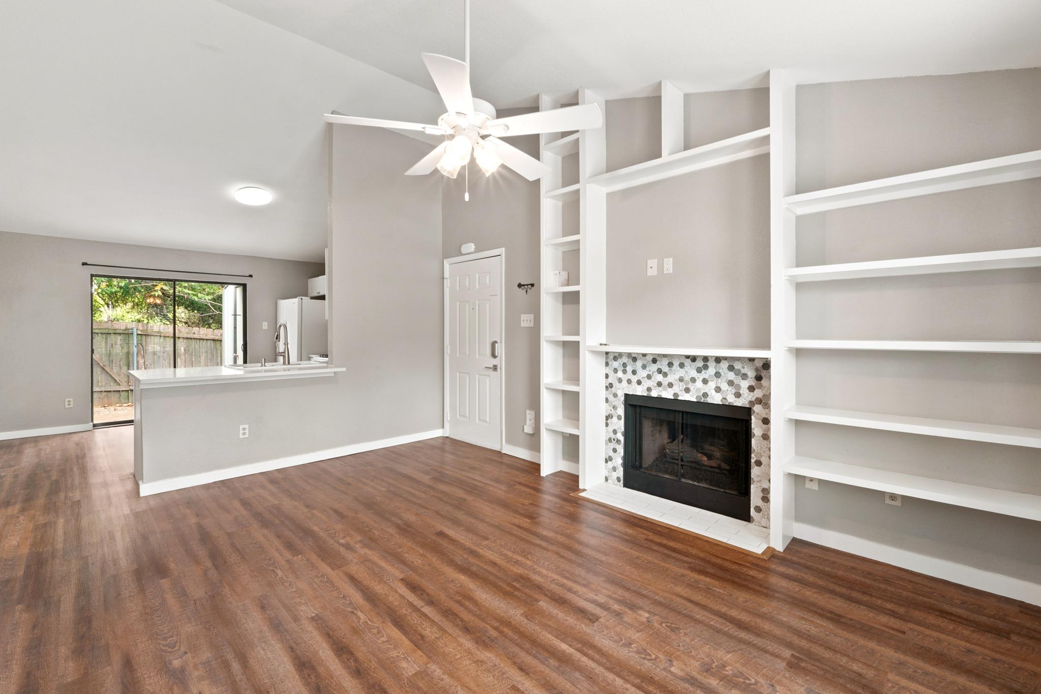 Empty living room with a tiled fireplace, white built-in shelves, wood-look floors, and kitchen pass-through.