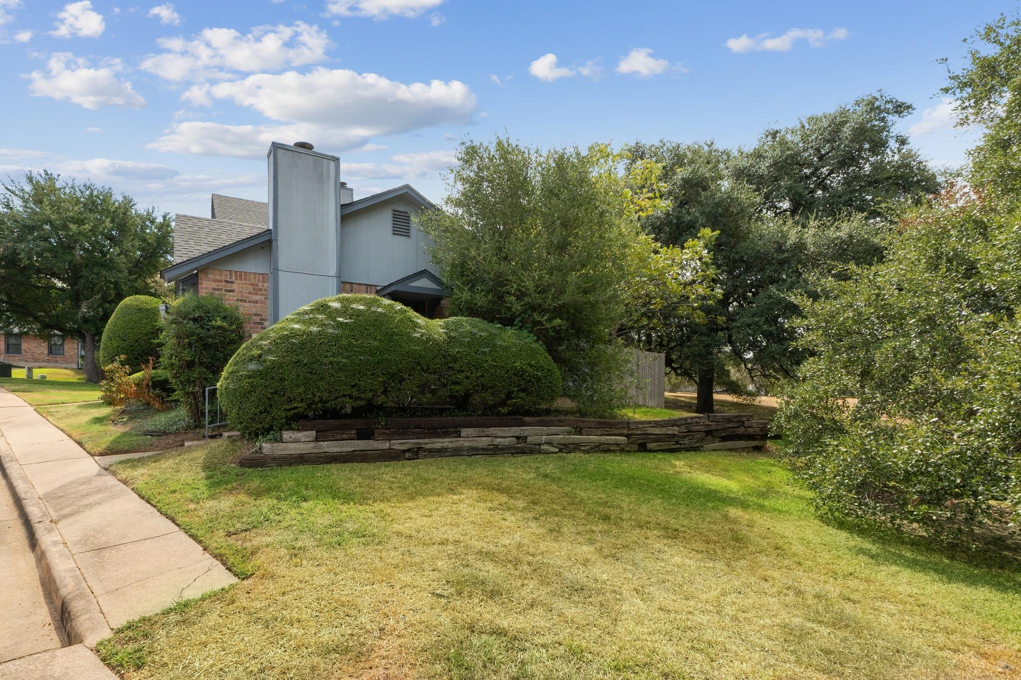Two-story townhouse with brick and gray siding, landscaped shrubs, trees, and a low timber retaining wall.