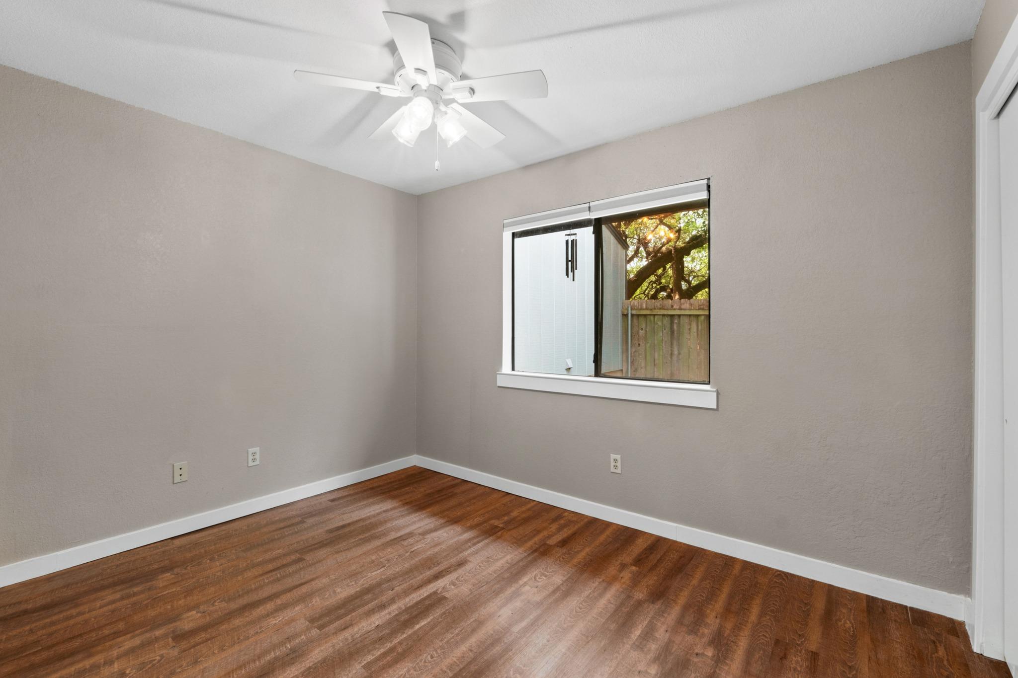 Empty bedroom with wood floors, a ceiling fan, and a window overlooking trees.