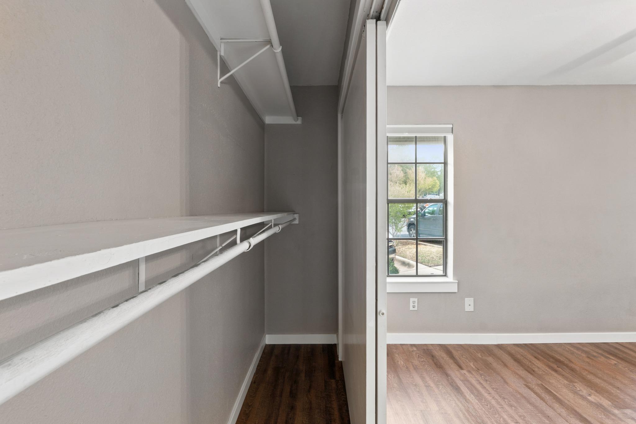 Empty walk-in closet with white shelves and hanging rods beside a bedroom window.