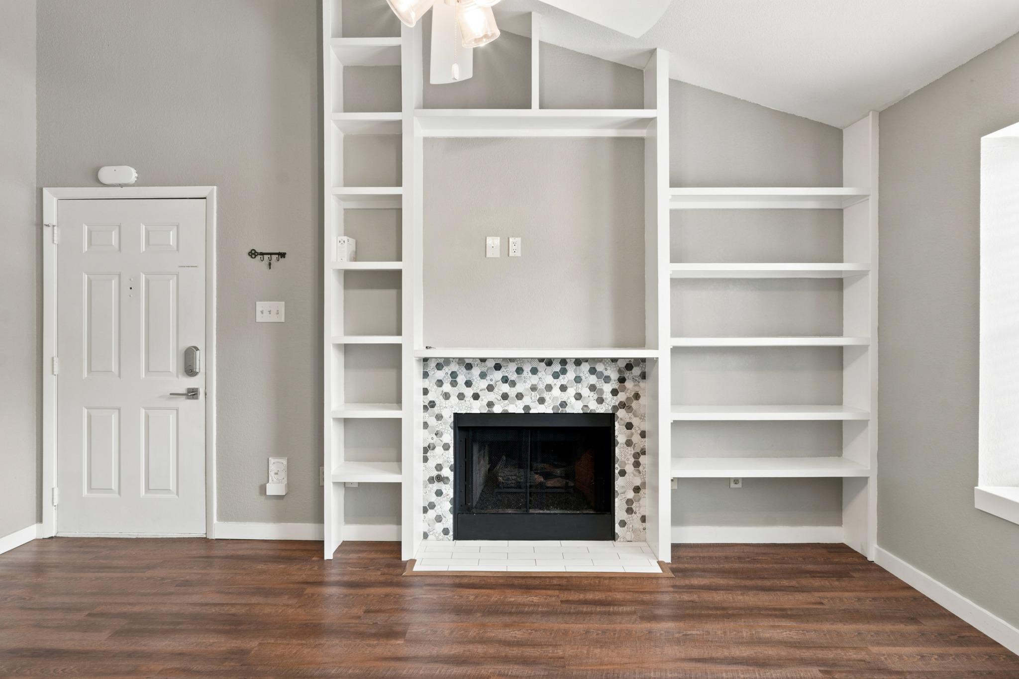 Vacant living room with built-in shelves framing a fireplace with a hexagonal tile surround.