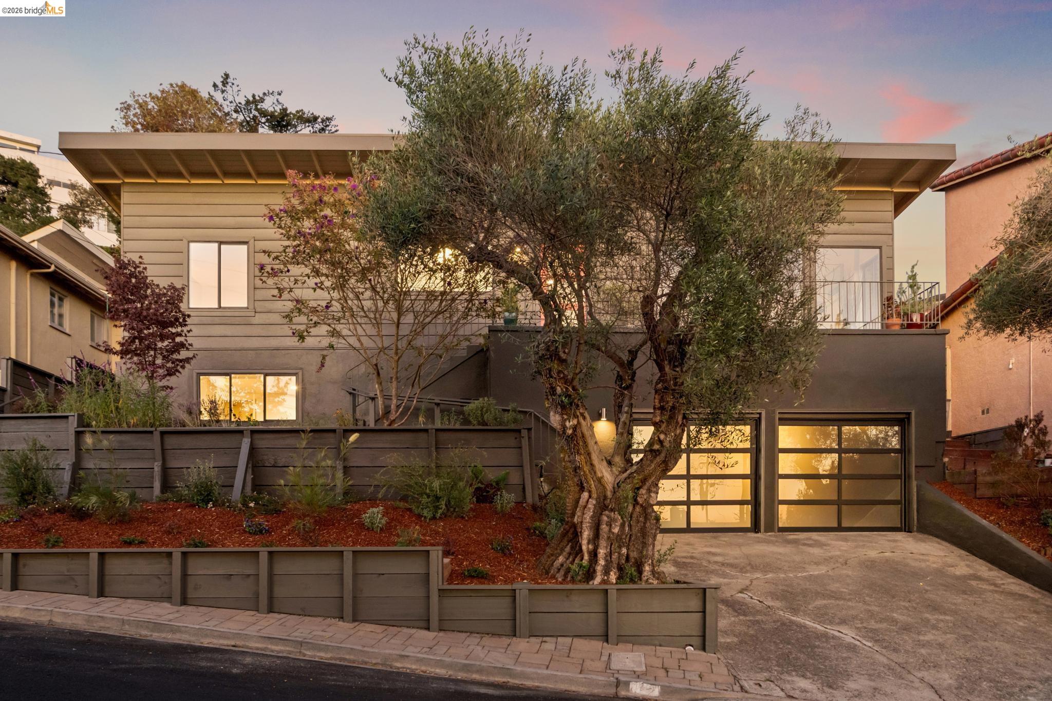 Modern two-story home with illuminated garage doors, an upper balcony, and a large olive tree in front.