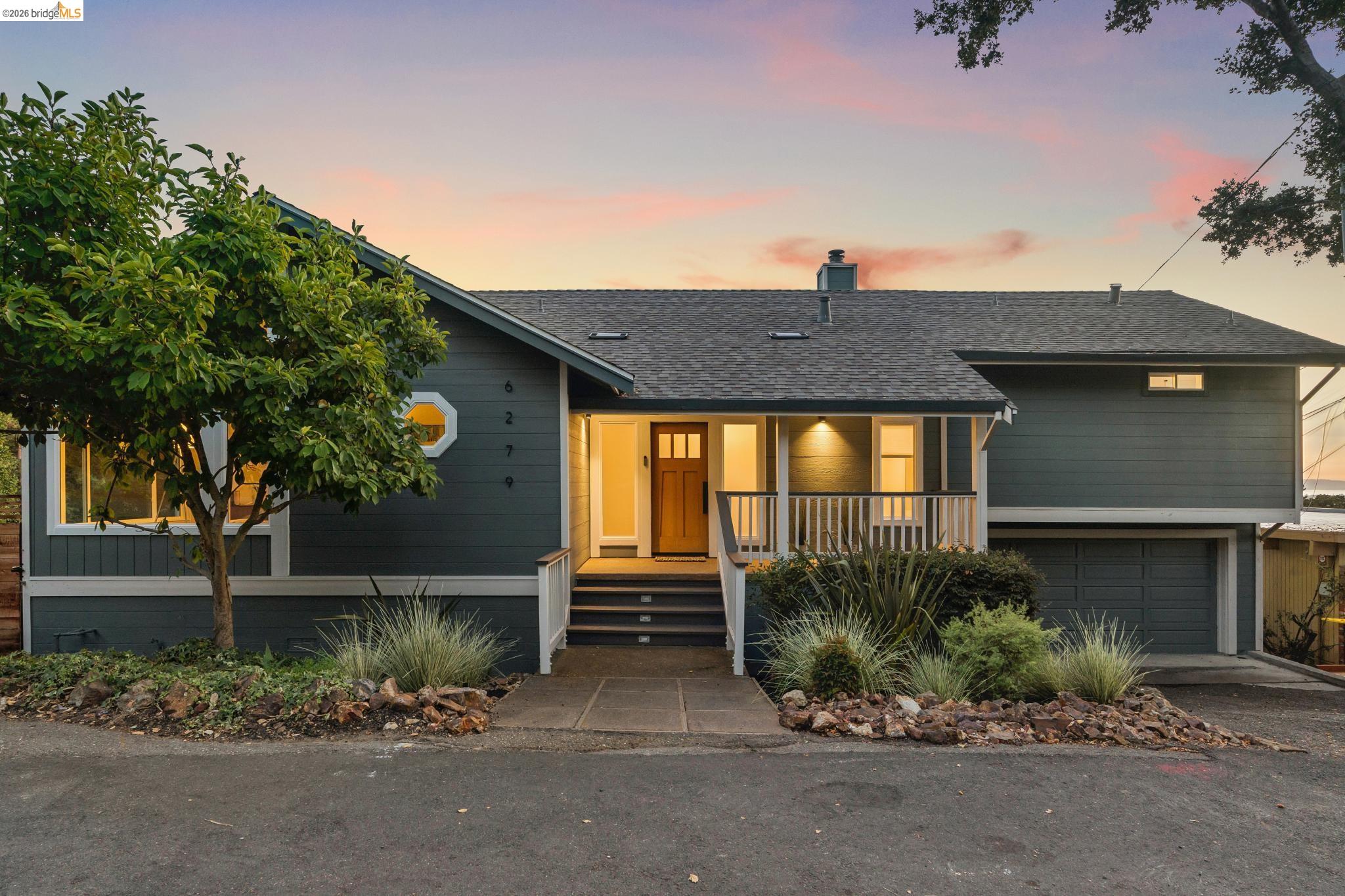 Dark gray house at dusk with a warmly lit front porch, attached garage, and landscaped entry.