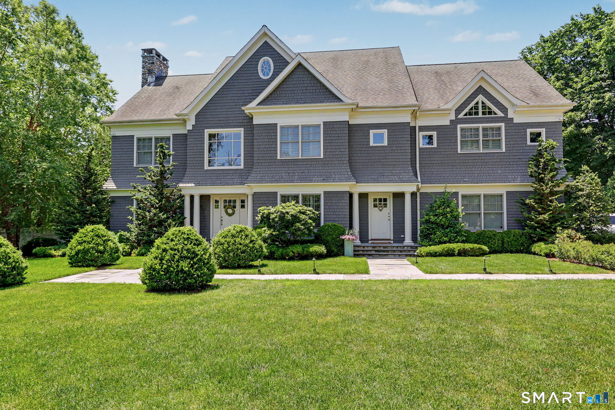 Gray-shingle house with white trim, multiple gables, landscaped shrubs, and a broad front lawn.