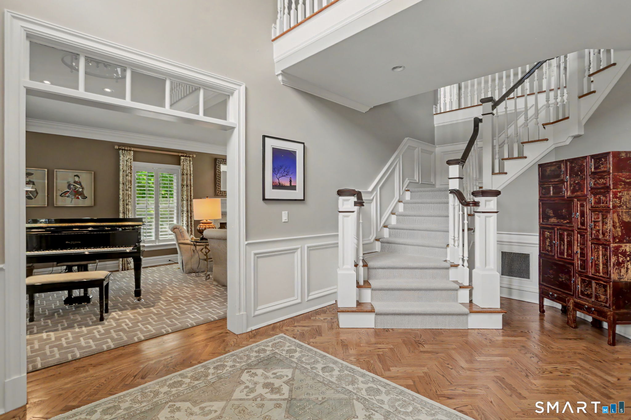Foyer with a carpeted staircase, herringbone wood floor, and adjoining sitting room with a grand piano.