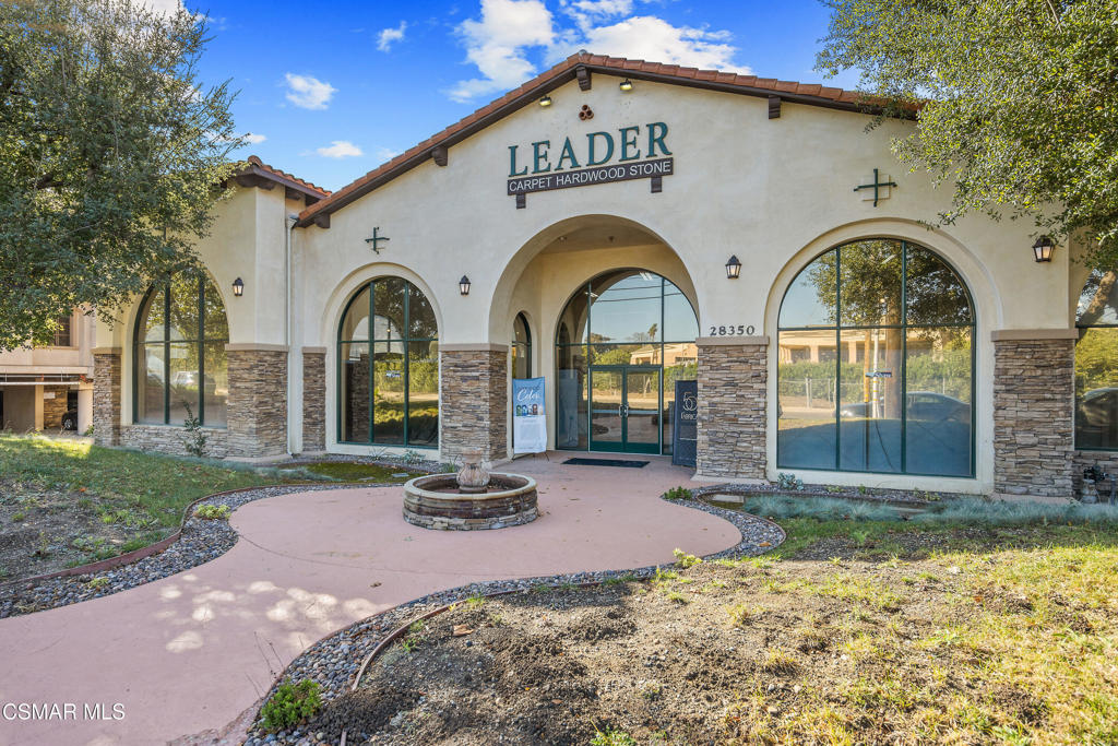 Commercial building with arched windows, stone accents, and a fountain outside the LEADER Carpet Hardwood Stone storefront.