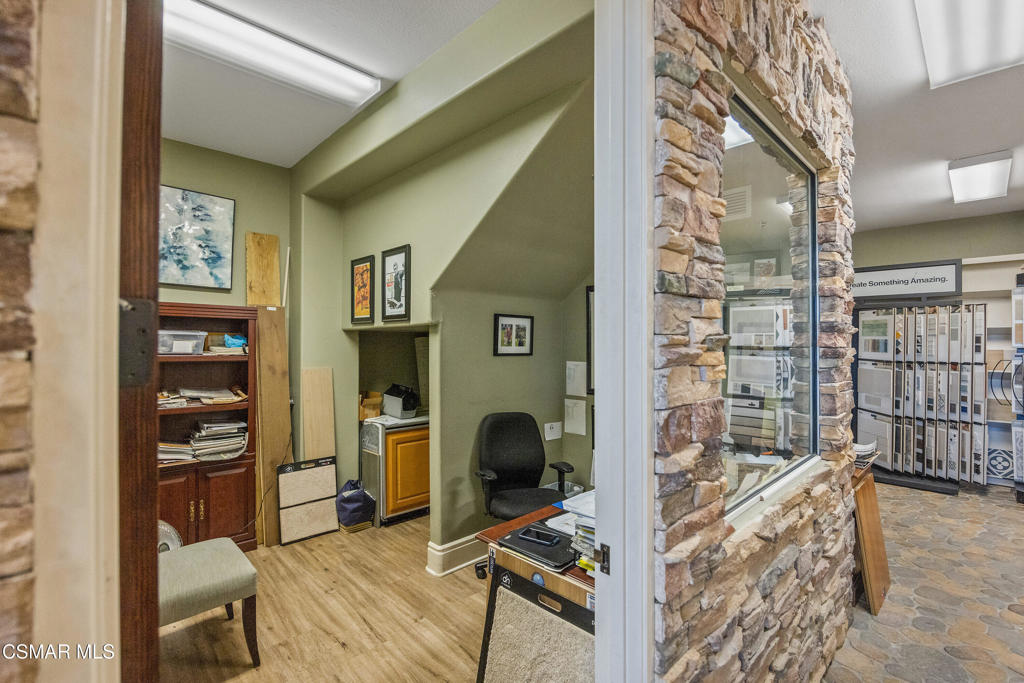 Office with wood shelving and desk beside a stone-framed glass partition and showroom.