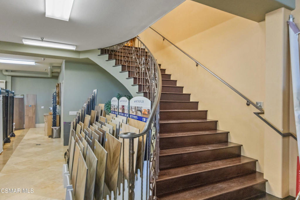 Showroom staircase with a curved wrought-iron railing beside rows of flooring samples.