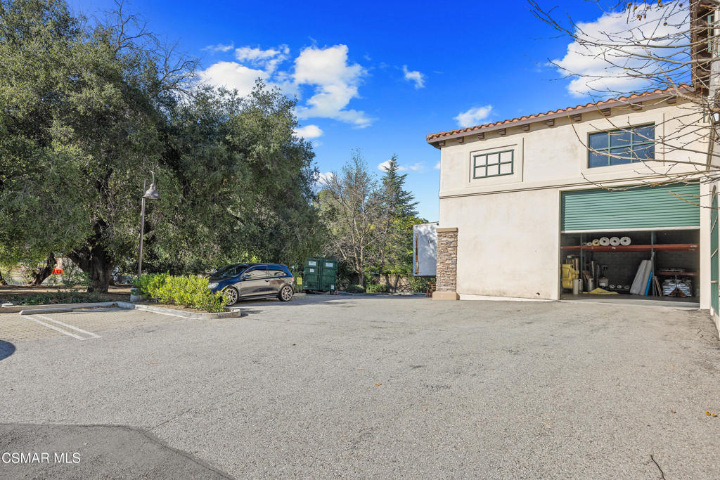 Paved parking area beside a stucco building with an open storage bay and mature trees.