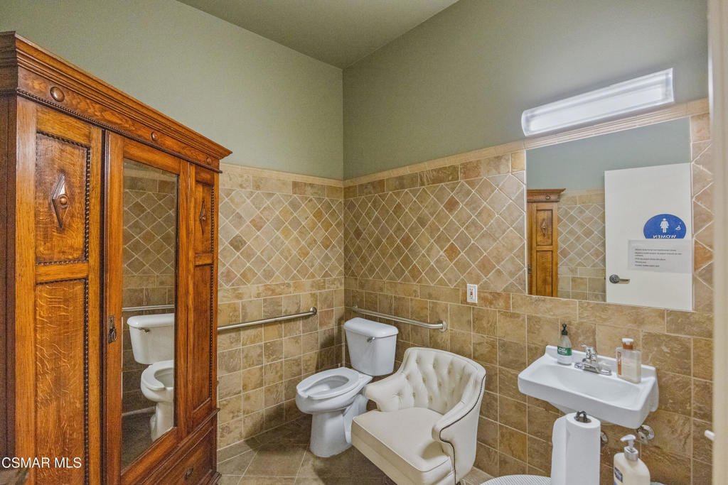 Tiled bathroom with a wooden mirrored cabinet, grab bars, and an upholstered chair beside the toilet.