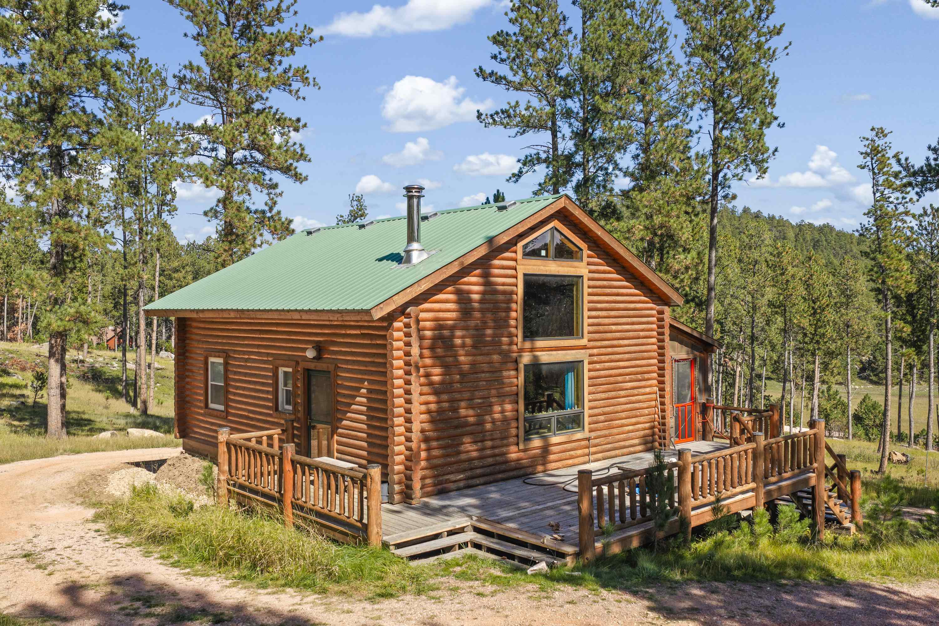 Log cabin with a green metal roof and wraparound deck among pine trees.