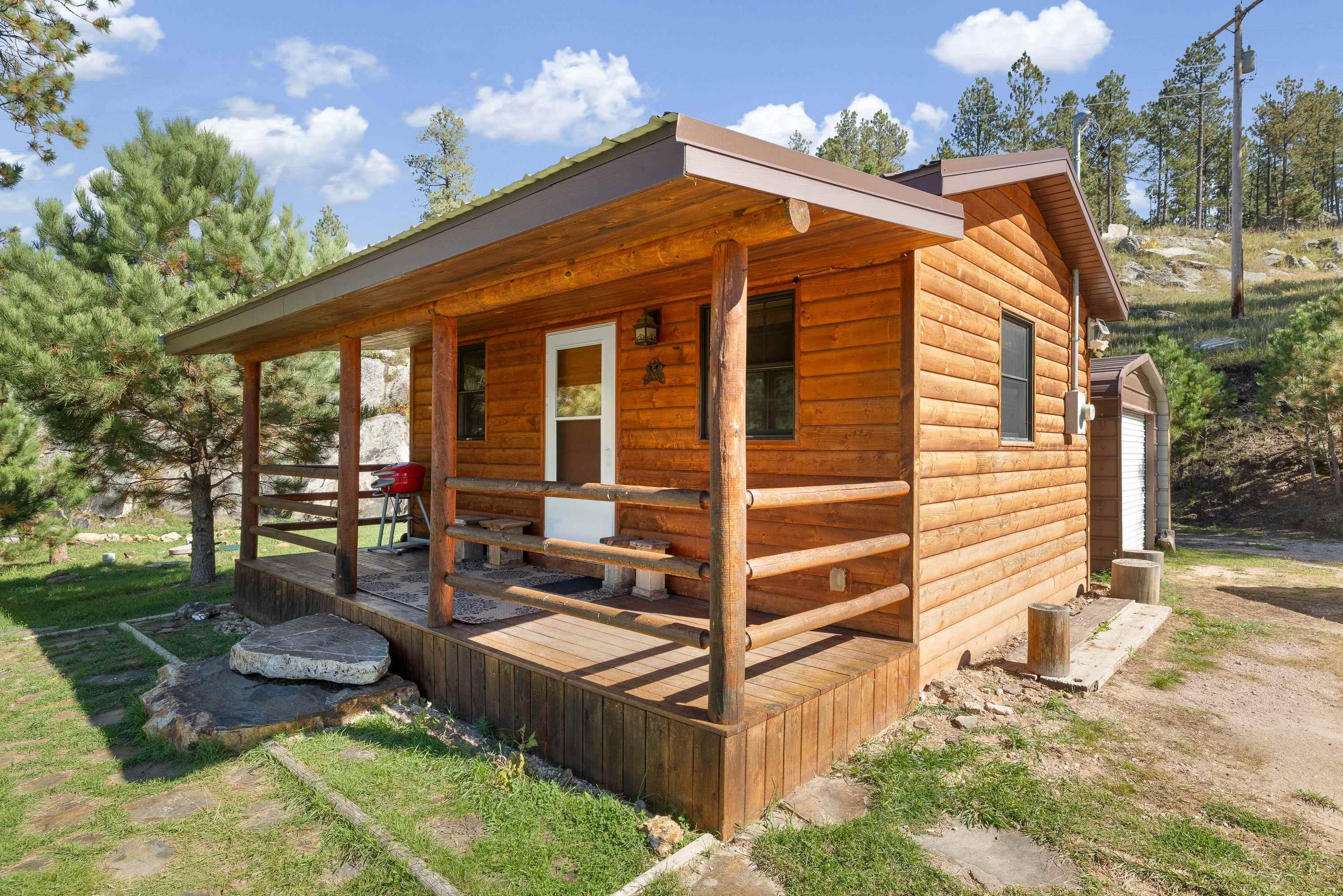 Rustic log cabin with a covered porch, surrounded by pine trees and rocky ground.