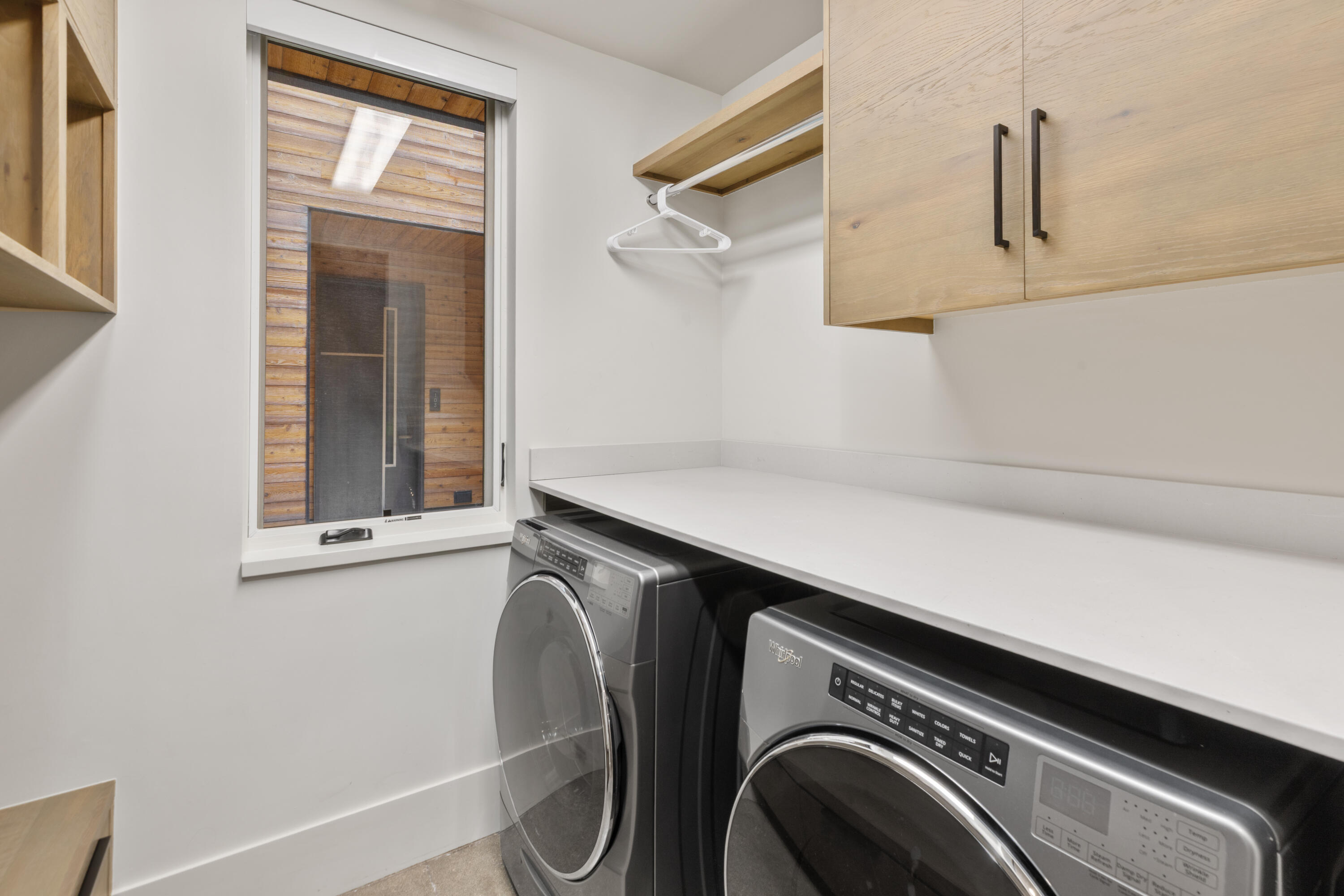 Laundry room with front-loading washer and dryer, white countertop, light wood cabinets, and a window.