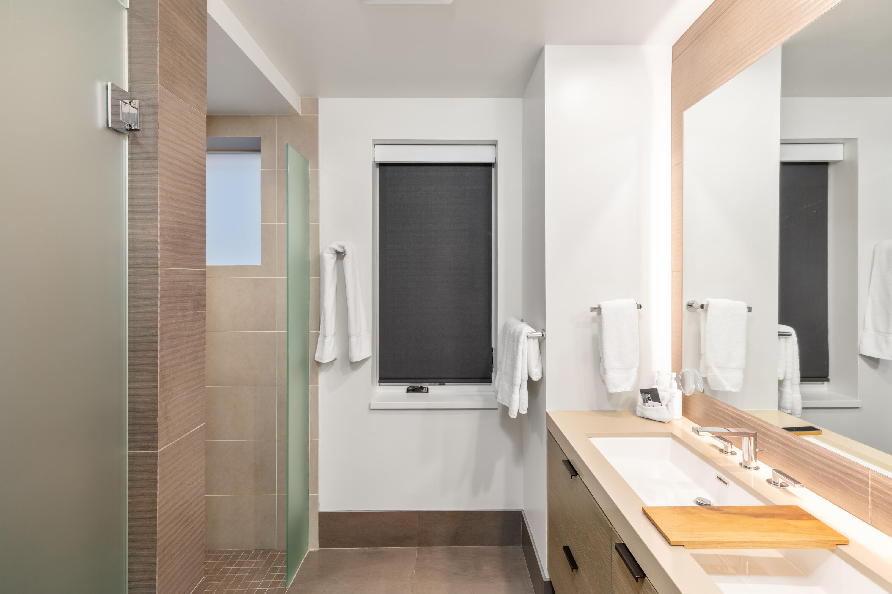 Modern bathroom with a glass shower, beige tile, and a long vanity beneath a large mirror.