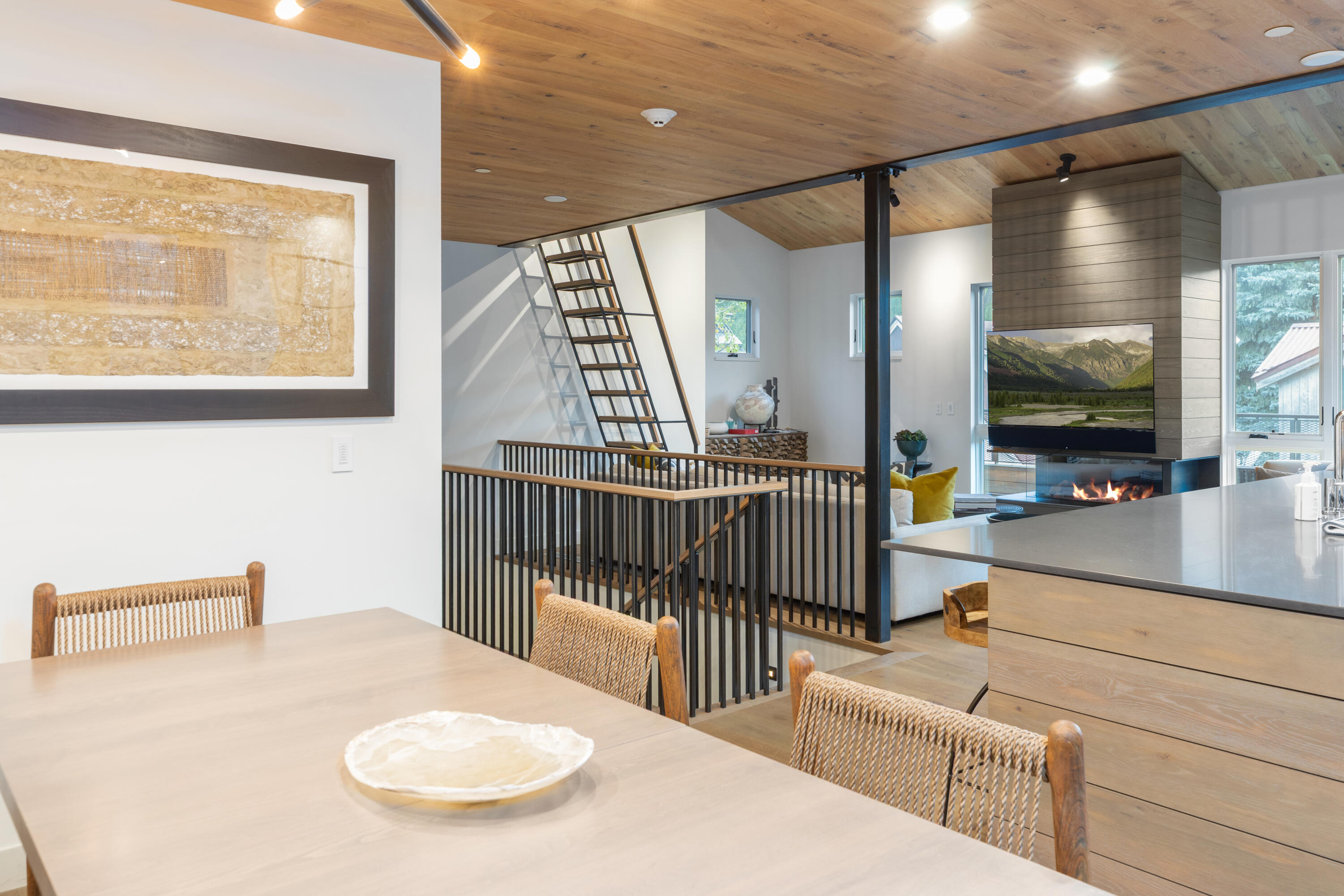 Open-plan dining and living area with a wood ceiling, black-railed stairwell, and modern fireplace.