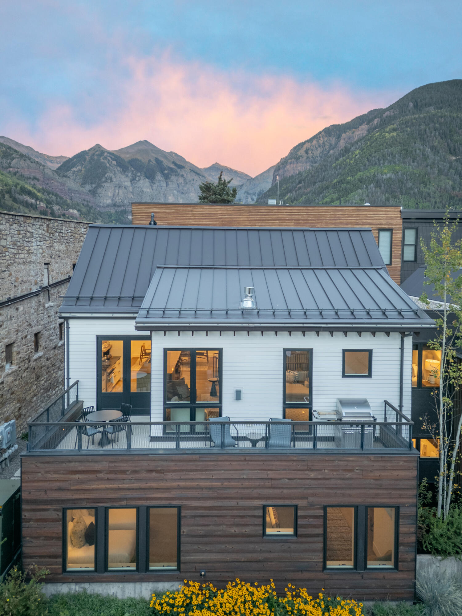 Modern Telluride home with a deck and metal roof, backed by mountains at sunset.