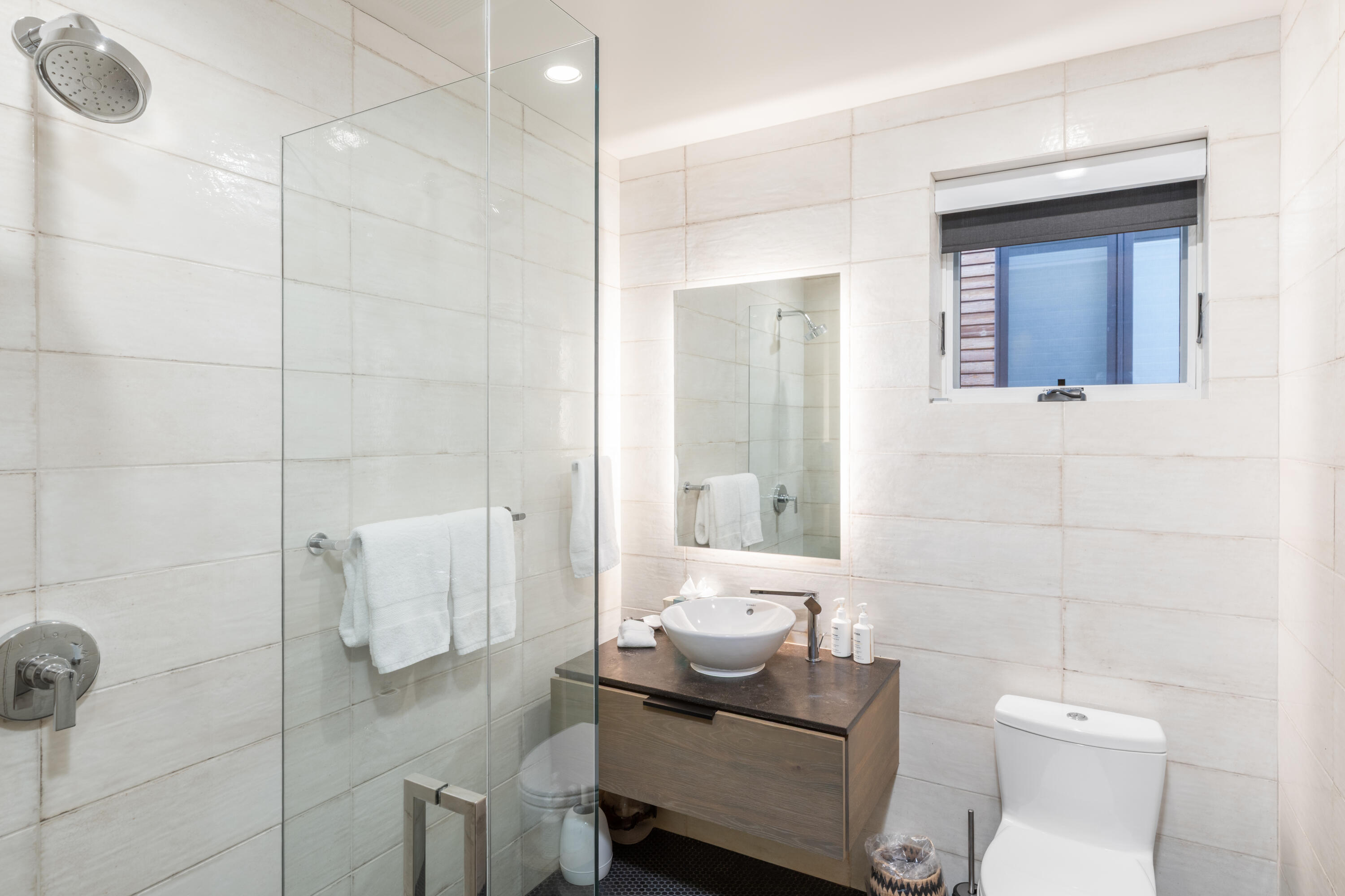 Modern tiled bathroom with a glass shower, floating wood vanity, vessel sink, and window.