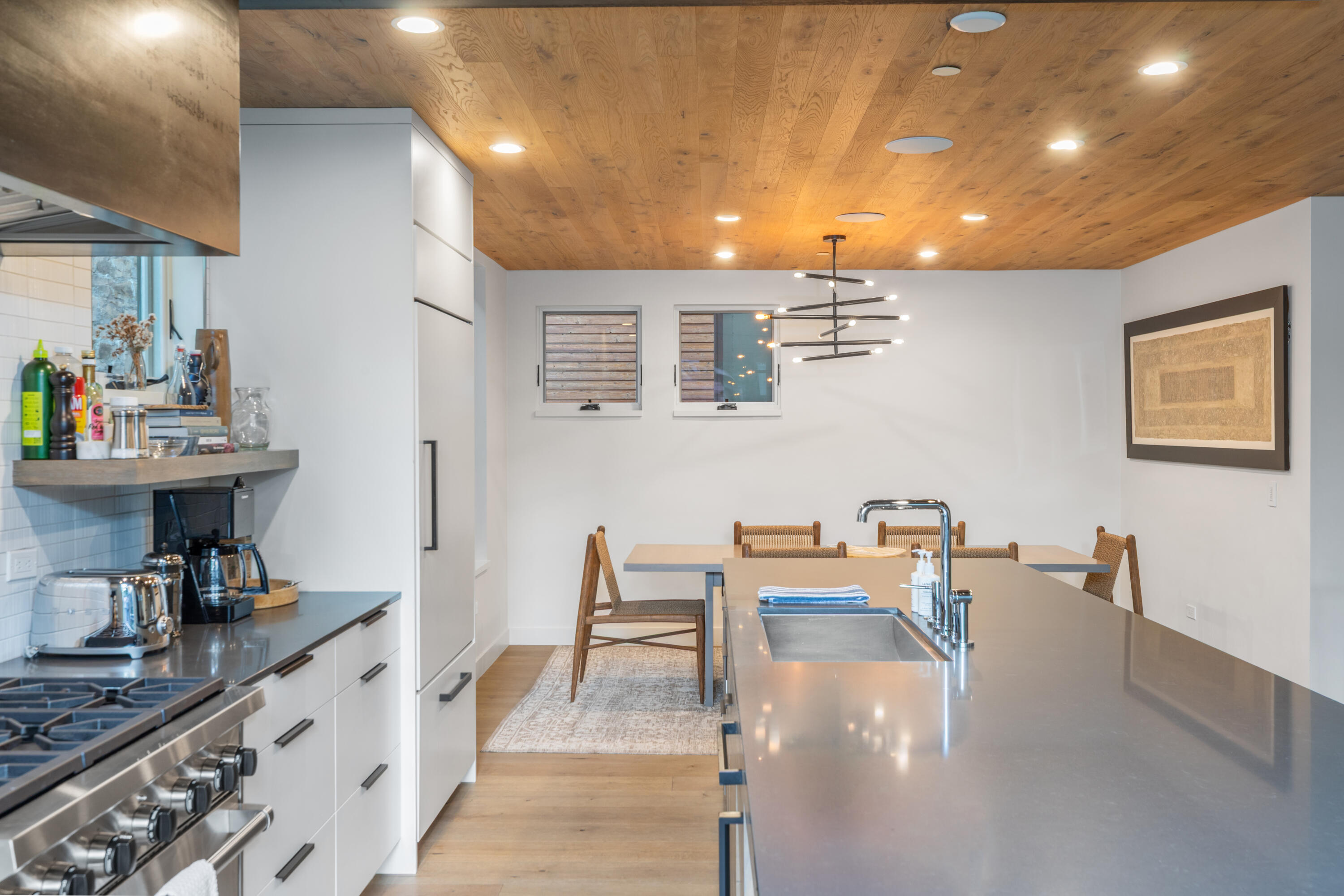 Modern kitchen with a large gray island, stainless-steel range, and dining table beneath a wood ceiling.