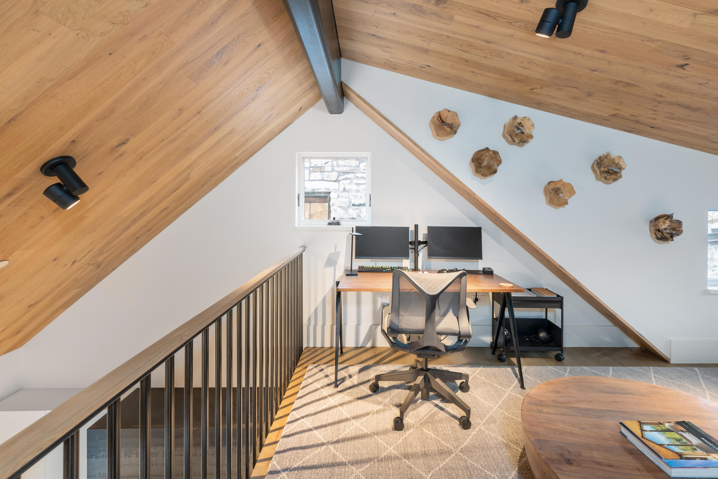 Loft office with dual monitors beneath a sloped wood ceiling and beside a black metal railing.
