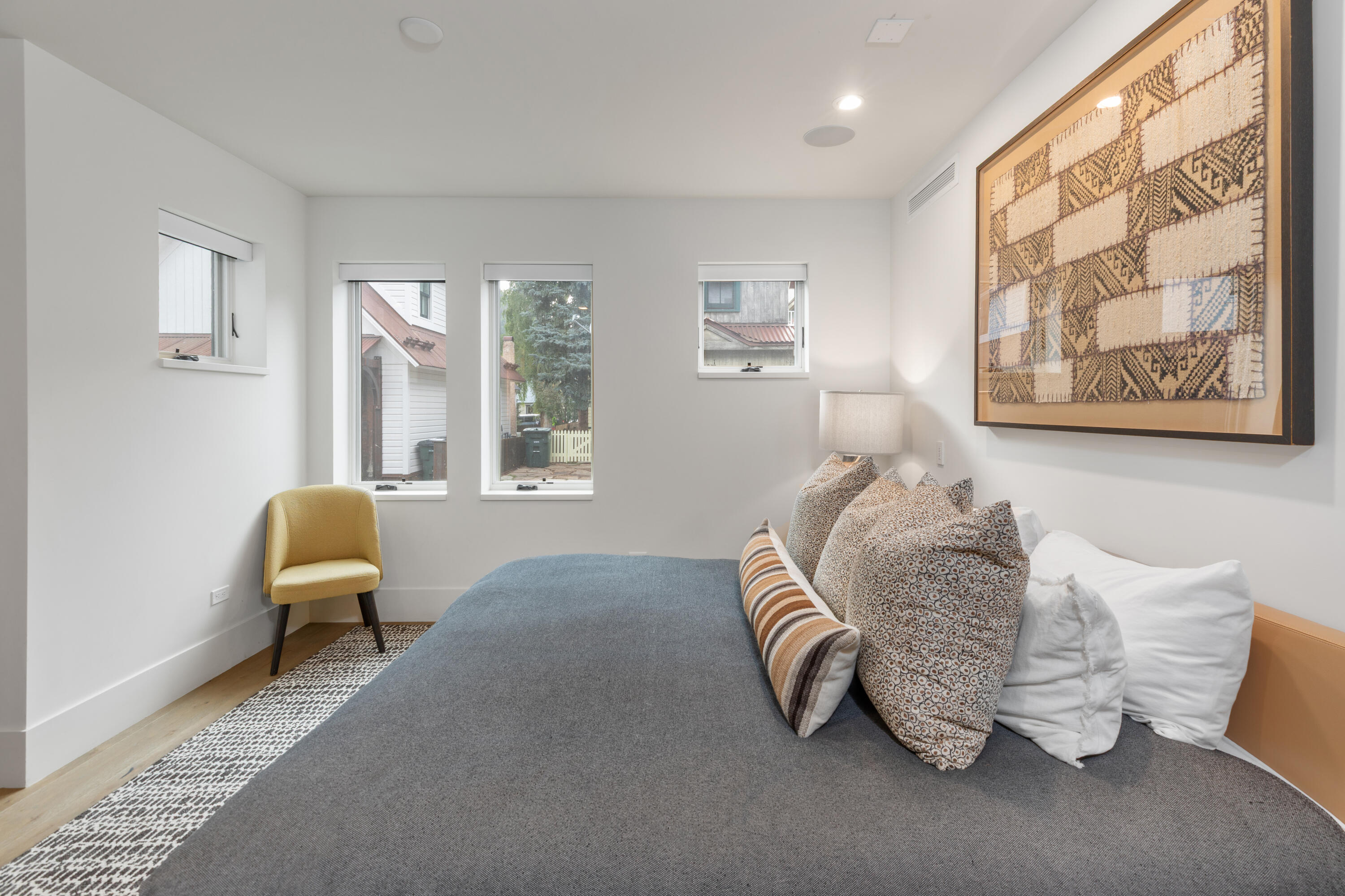 Bedroom with a gray-covered bed, patterned pillows, mustard-yellow chair, and large framed textile artwork.