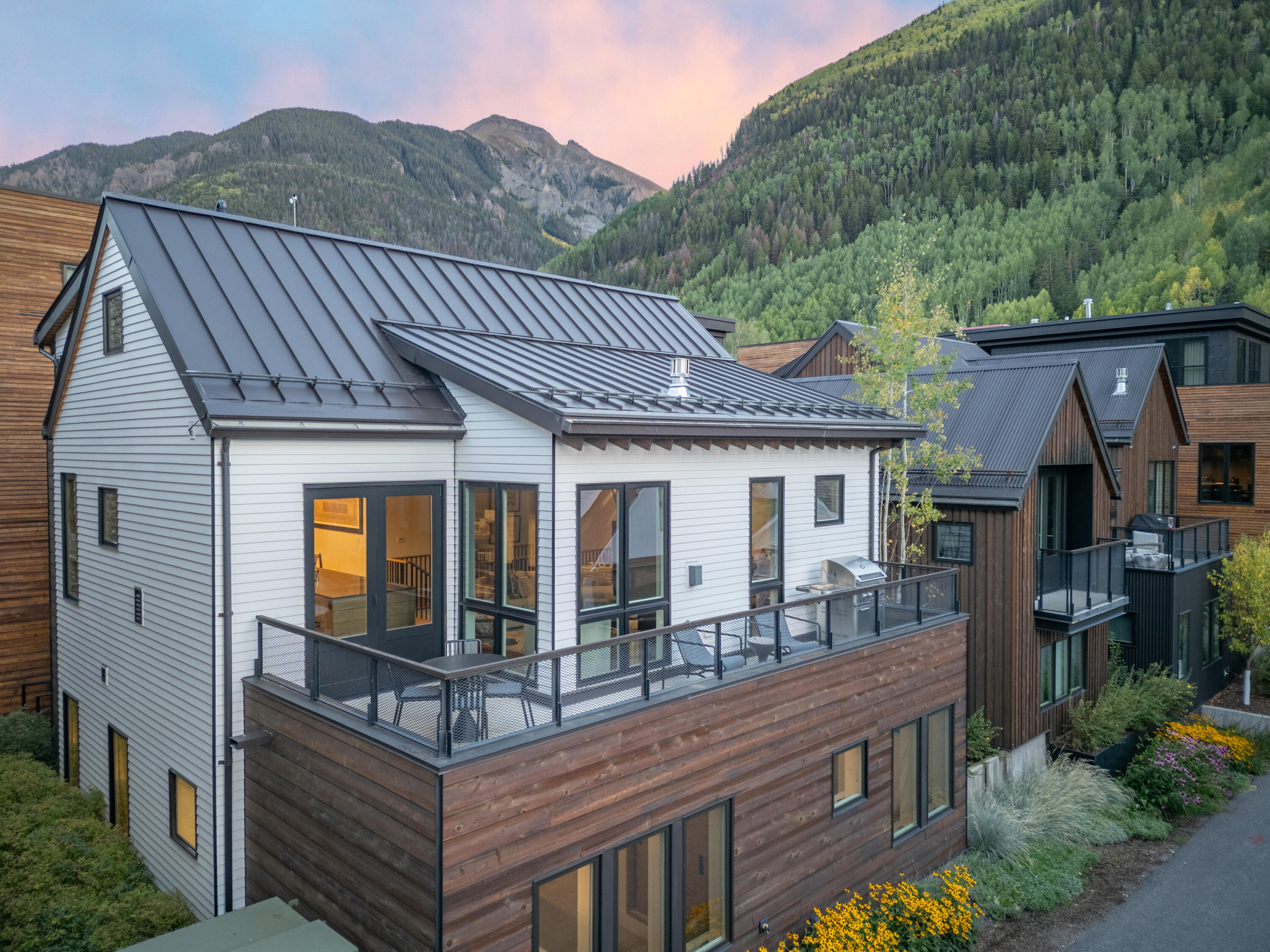 Modern mountain home with a wood-clad balcony, dark metal roofs, and forested mountains behind it.