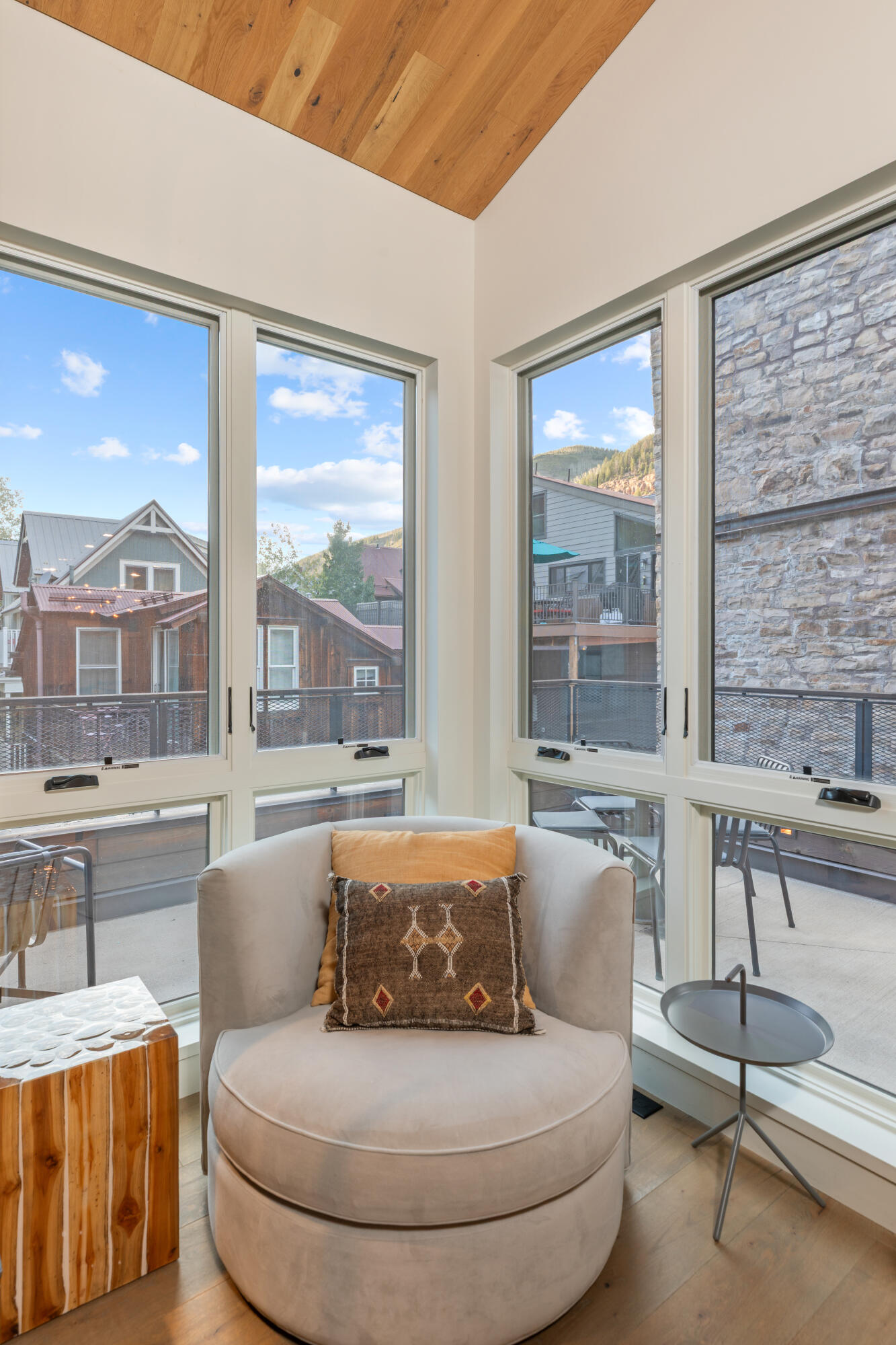 Sitting nook with a rounded cream chair, wood ceiling, and tall windows overlooking nearby buildings and a hillside.