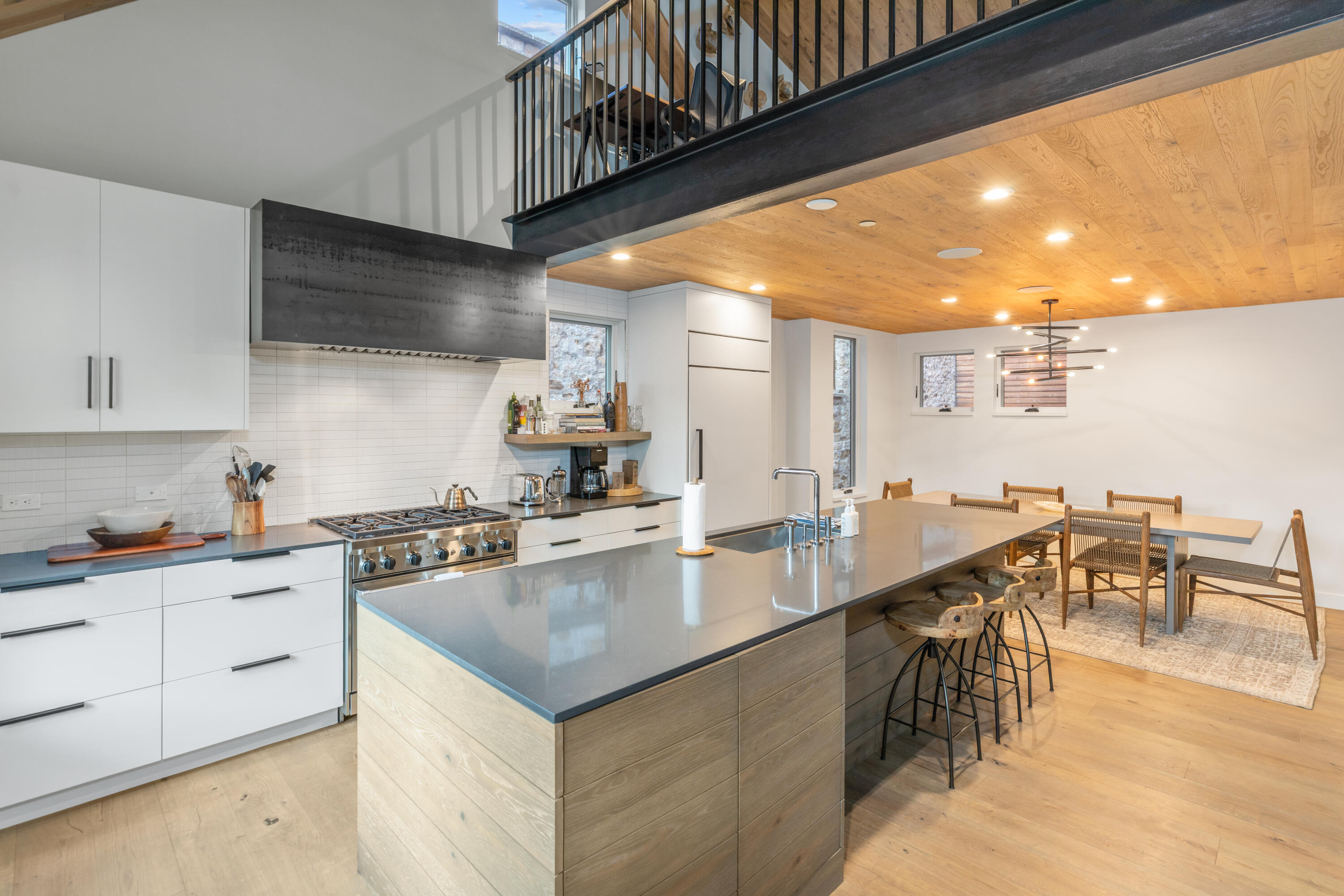 Modern kitchen with a large gray island, stainless steel range, wood ceiling, and dining area beneath a mezzanine.