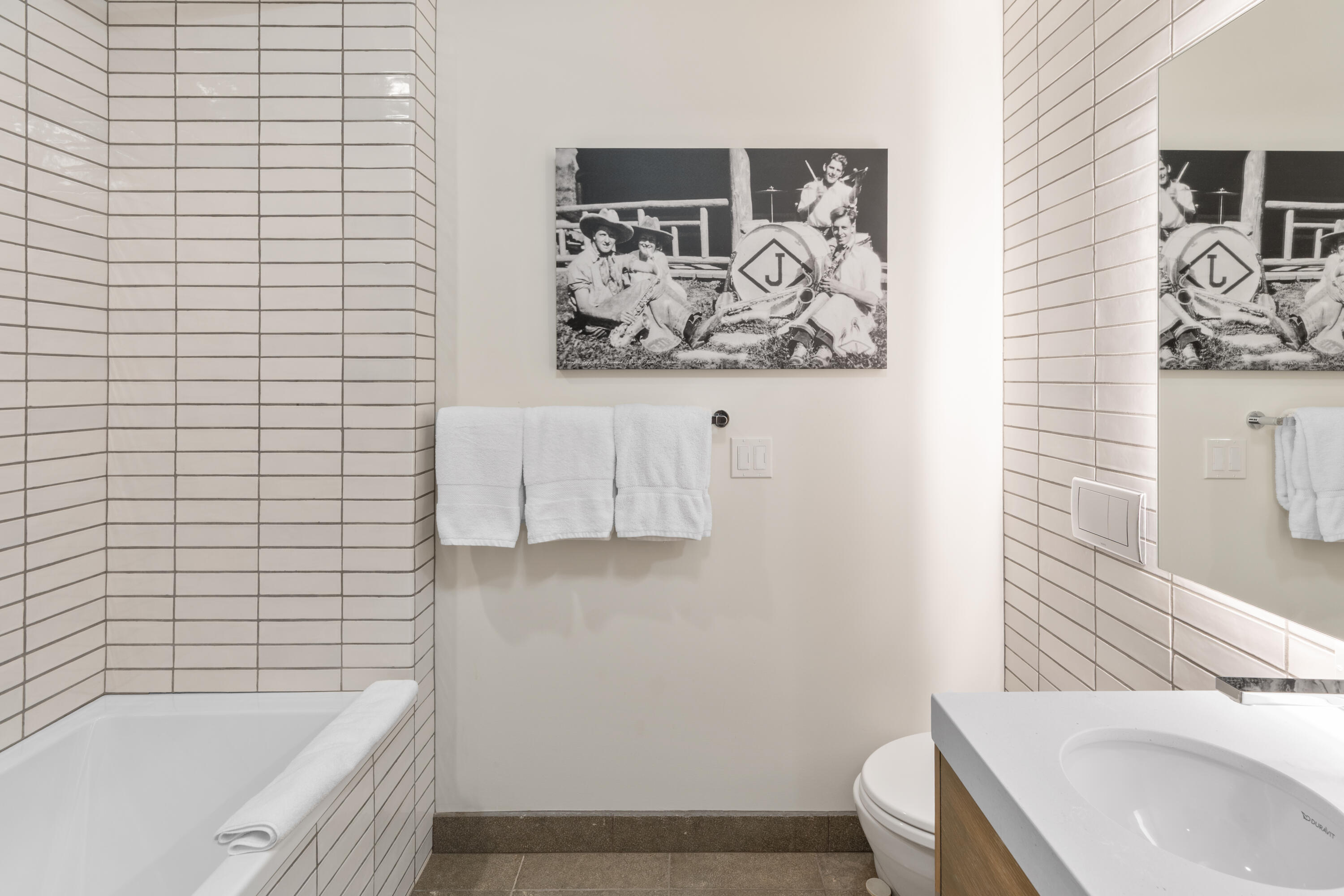 Bathroom with white tiled bathtub, towel bar, wood-front vanity, and framed black-and-white photograph.