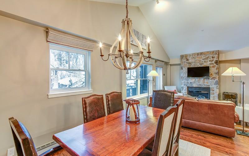 Dining area with a wooden table and chandelier beside a living room with a stone fireplace.