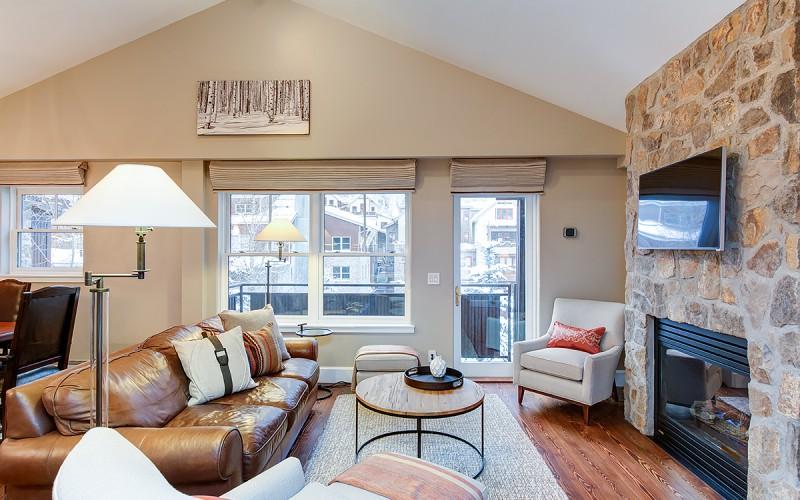 Living room with a vaulted ceiling, leather sofa, and stone fireplace beside large windows.