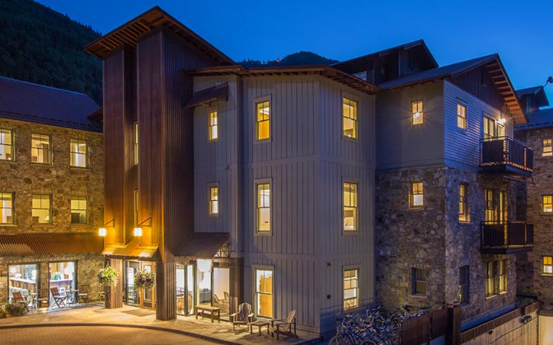 Multi-story building with stone walls, gray siding, balconies, and warmly lit windows at dusk.