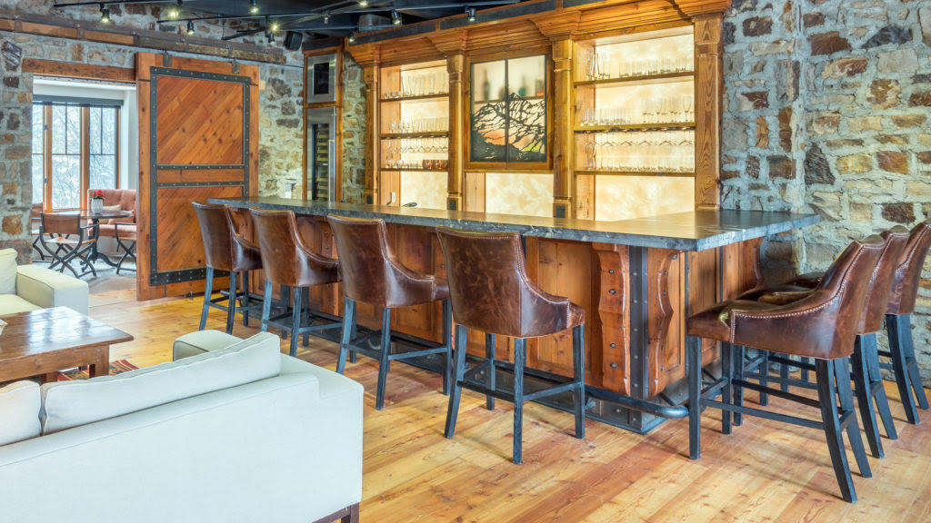 Interior bar with leather stools, illuminated glass shelves, stone walls, and a white sofa.