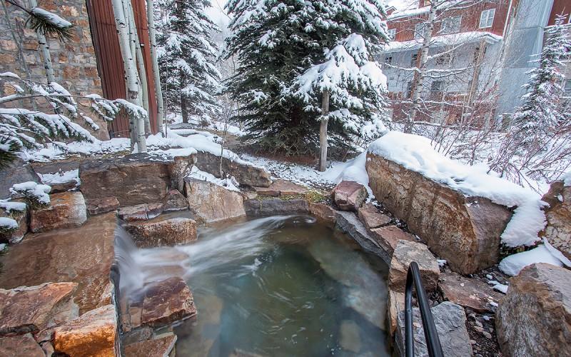 Outdoor hot tub bordered by snow-covered stonework and evergreen trees.
