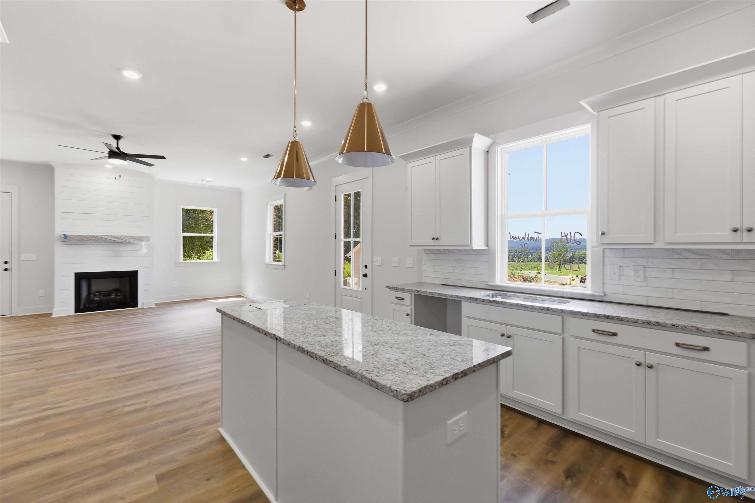 Bright vacant kitchen with granite island, white cabinetry, wood flooring, and an open living area with fireplace.