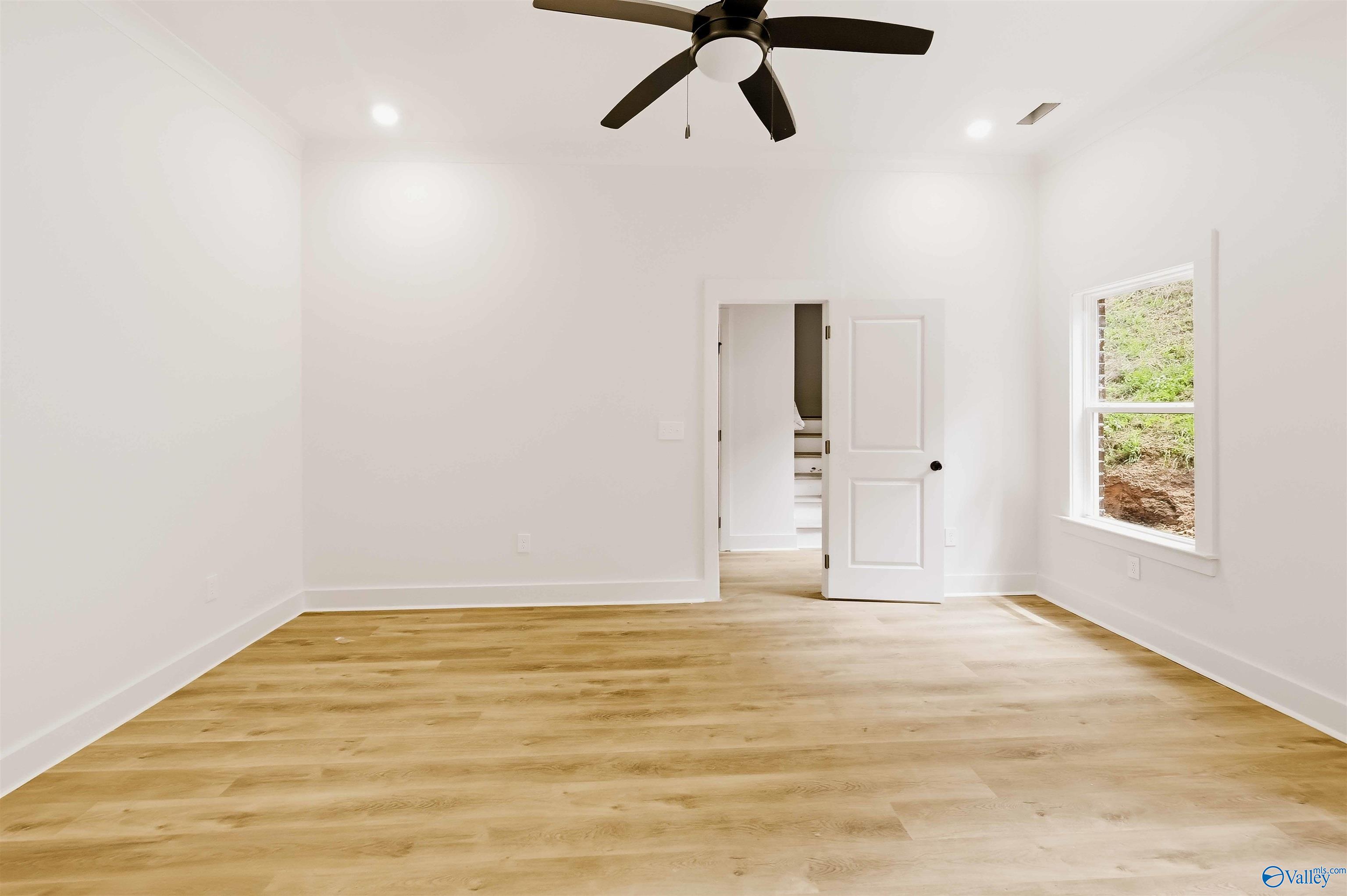 Empty room with light wood floors, a black ceiling fan, and a window overlooking greenery.