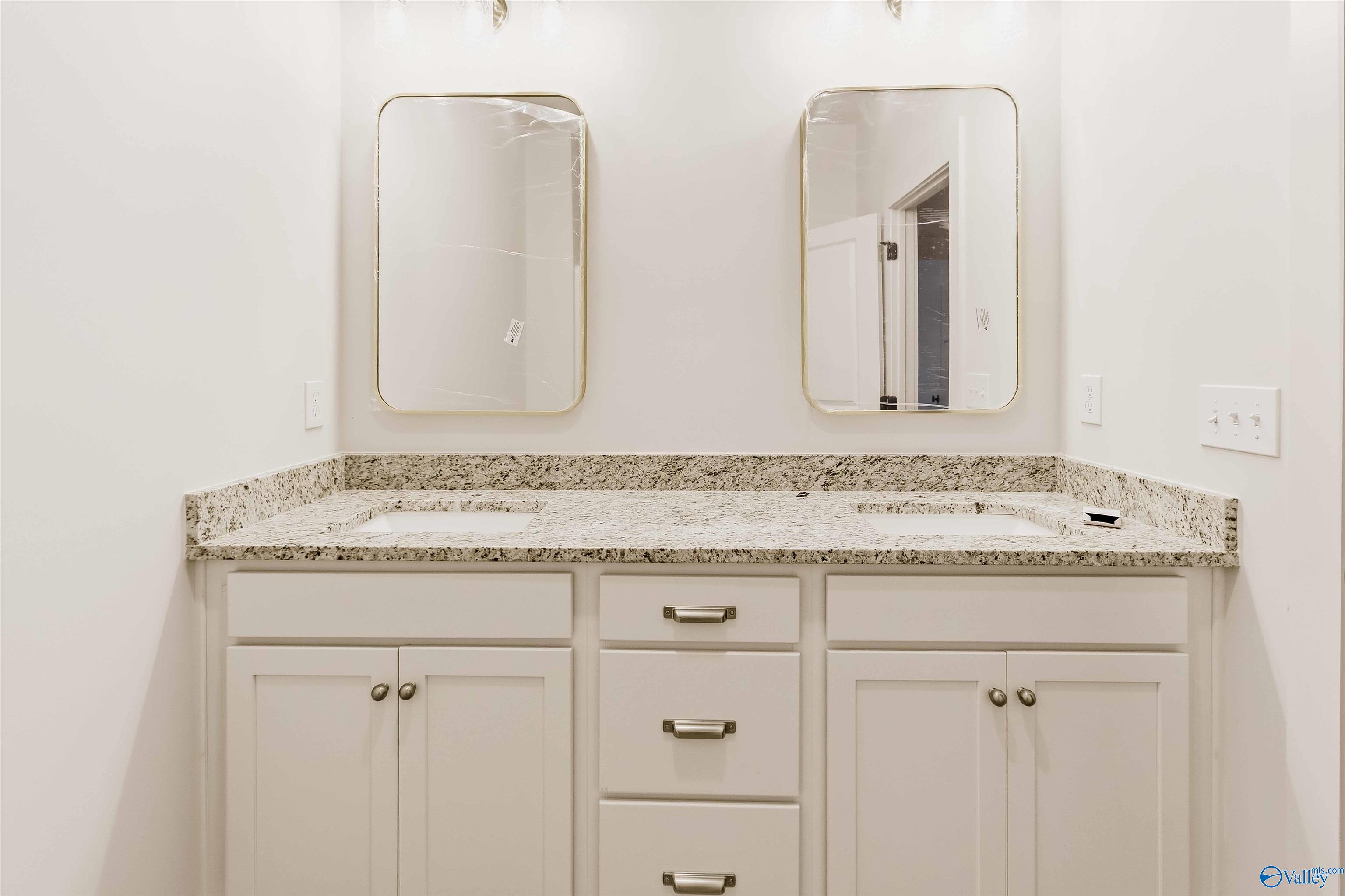 Bathroom vanity with two sinks, speckled granite countertop, white cabinetry, and gold-framed mirrors.