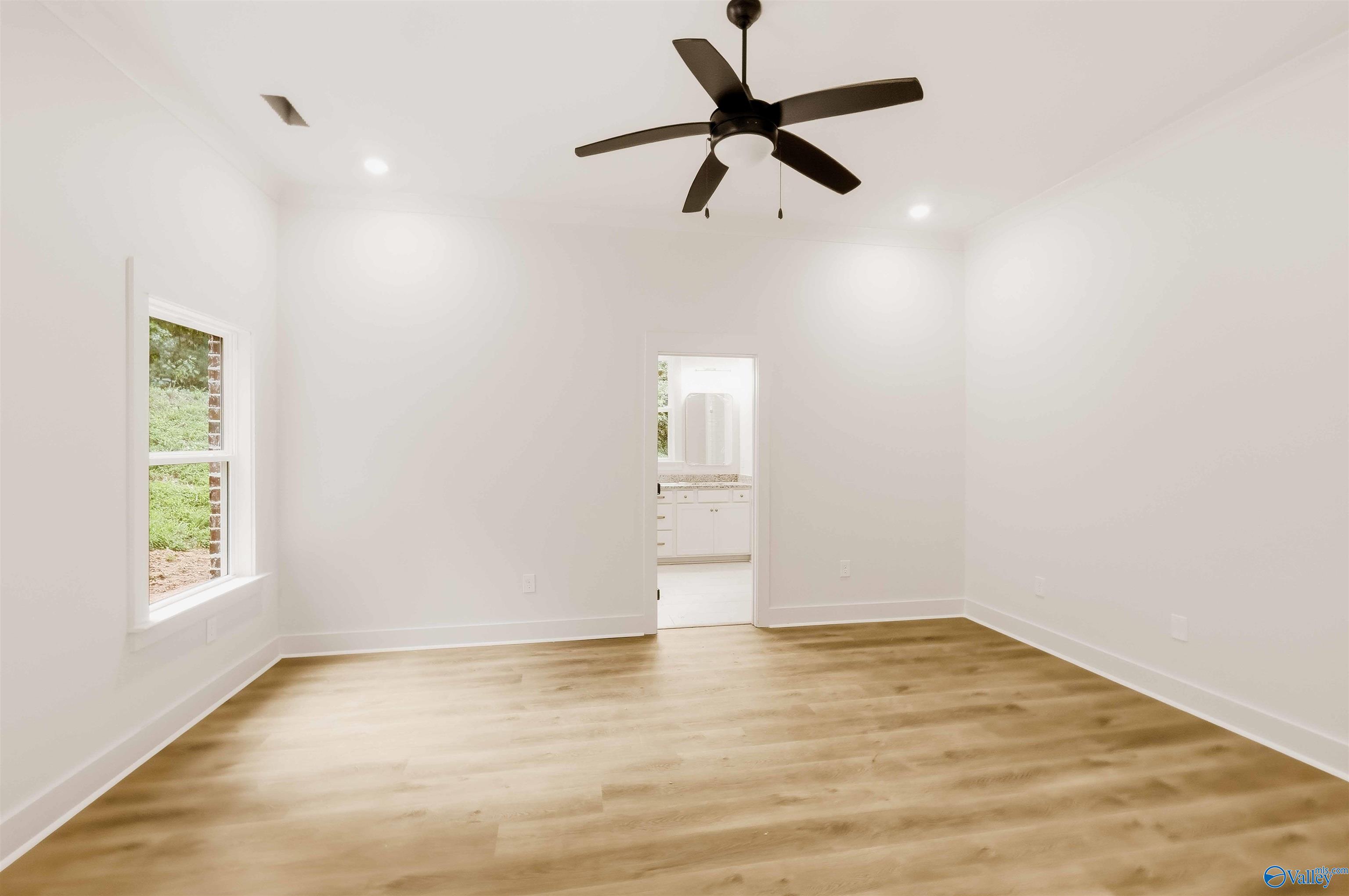 Empty room with light wood floors, a dark ceiling fan, and a doorway to a bathroom.