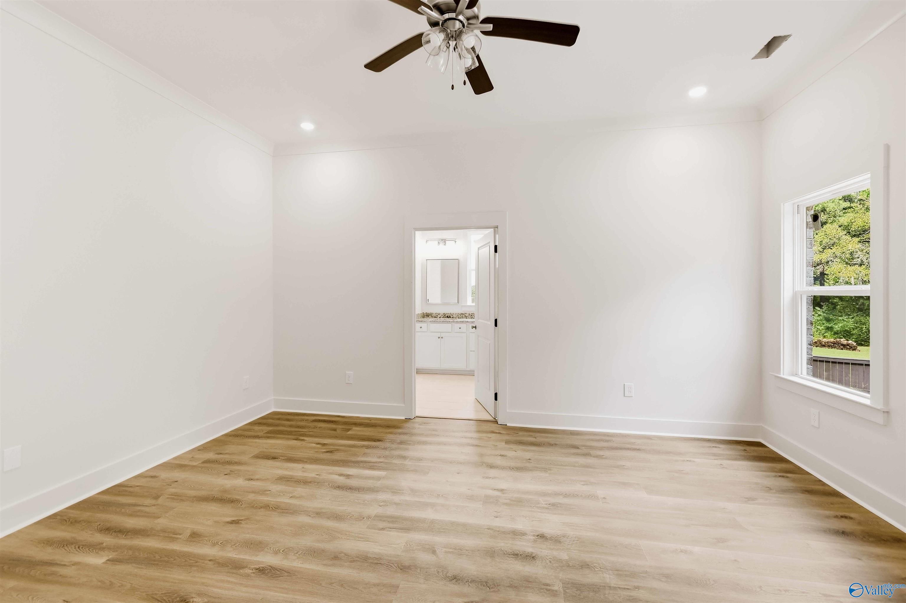 Vacant bedroom with wood-look floors, ceiling fan, and window overlooking greenery; bathroom vanity visible through doorway.