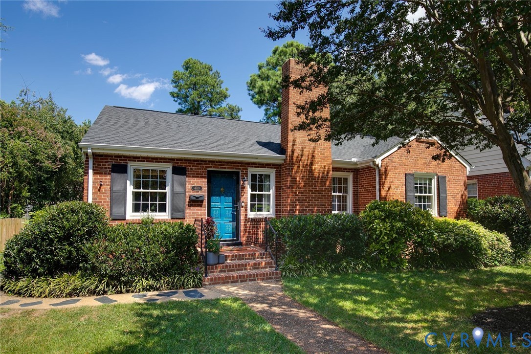 Red-brick ranch home with a bright blue front door, tall chimney, and landscaped shrubs.