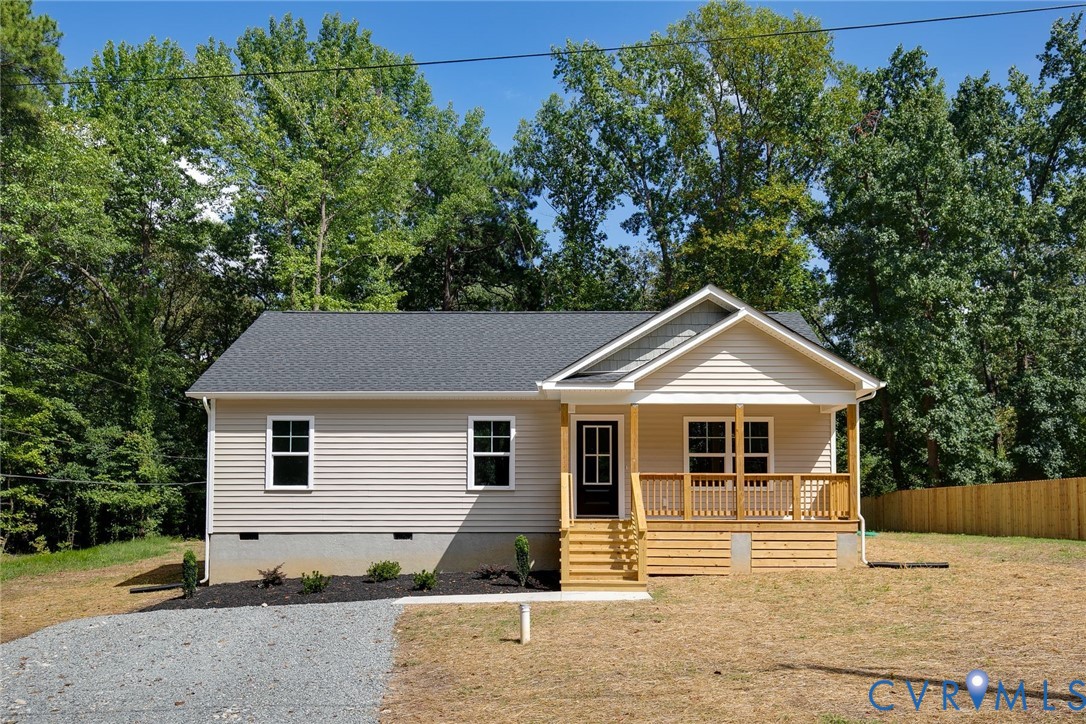 Beige single-story house with a covered wooden porch, gravel driveway, and trees behind it.