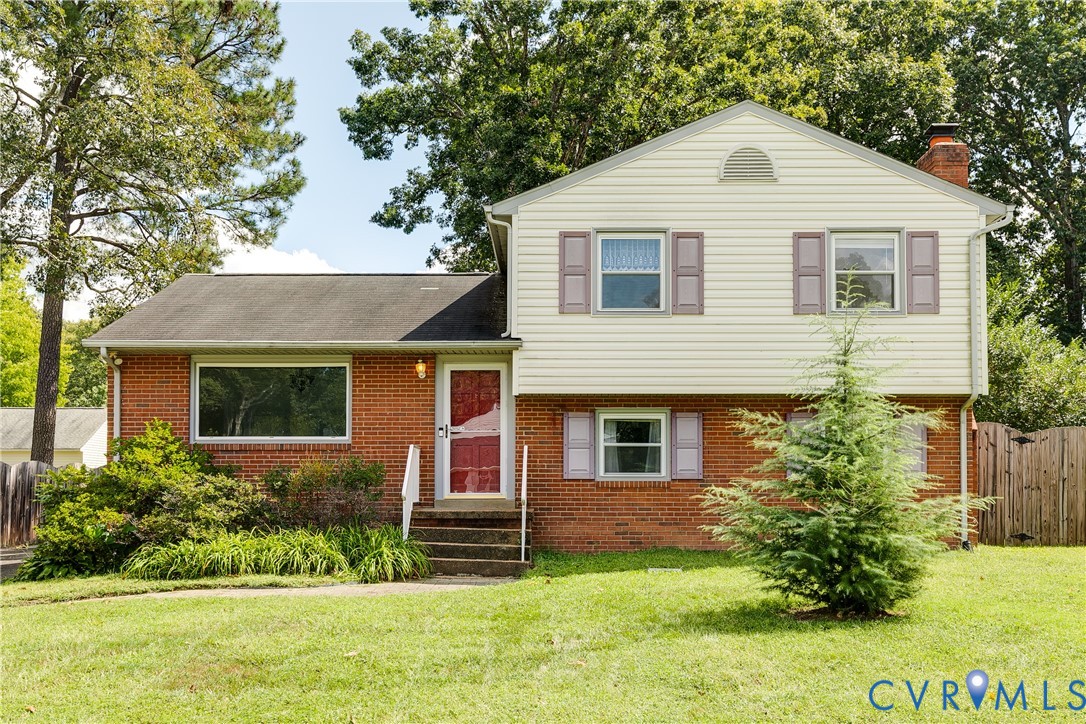 Split-level brick and siding house with a red front door, mauve shutters, and a tree-lined lawn.